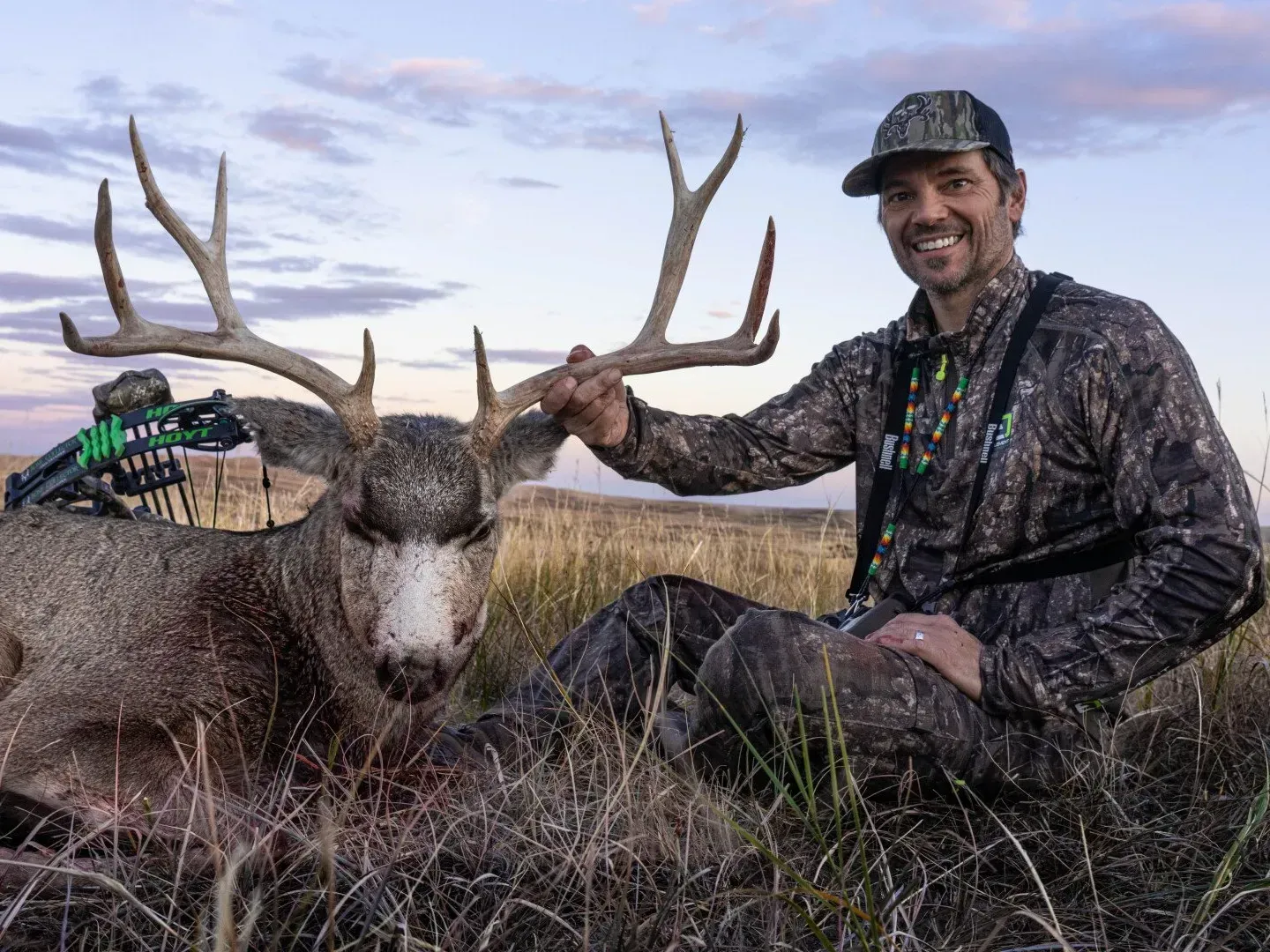 Man in camo kneels beside a buck he shot with a bow, grassy field, smiles.