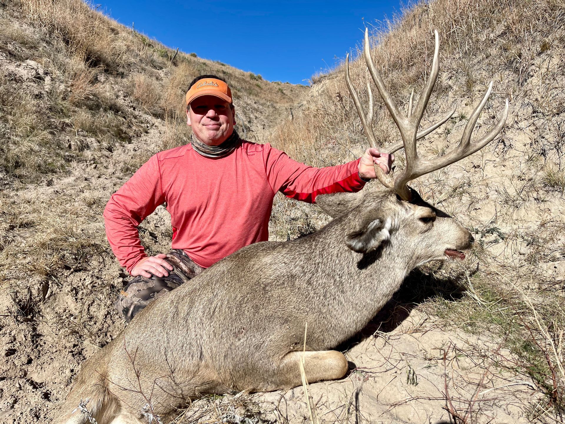 Man in orange shirt poses with a large buck in a hilly, outdoor setting.