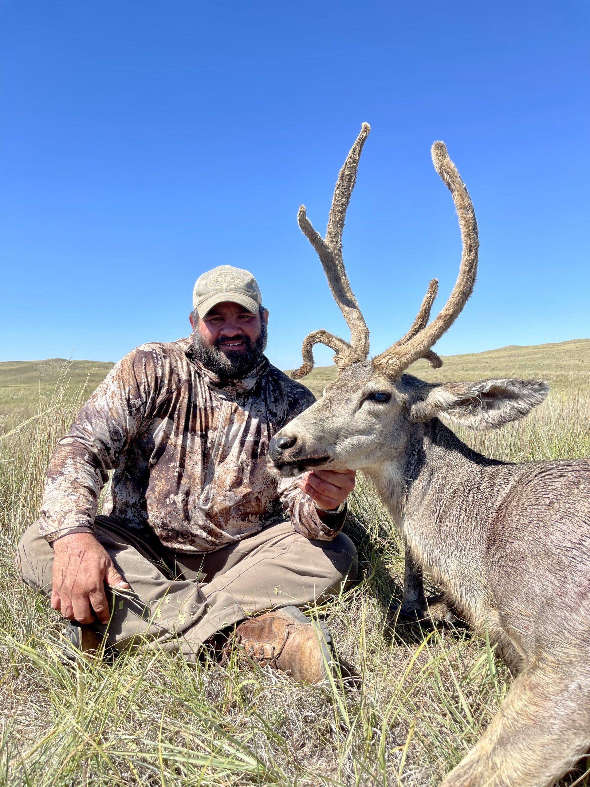 Man in camouflage with a harvested deer with large antlers in a grassy field under a blue sky.