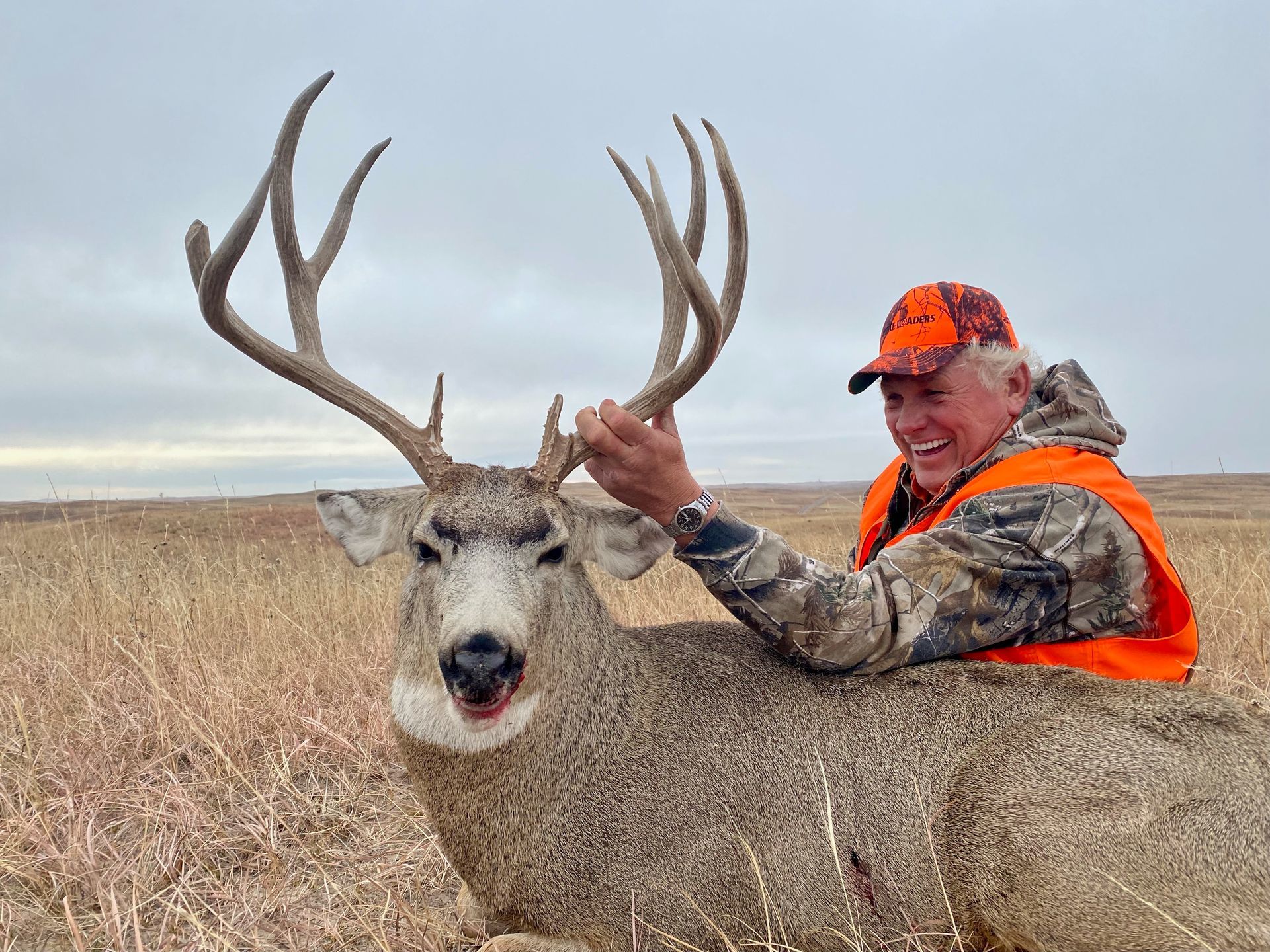 Hunter in orange gear smiles next to a large buck in a grassy field.