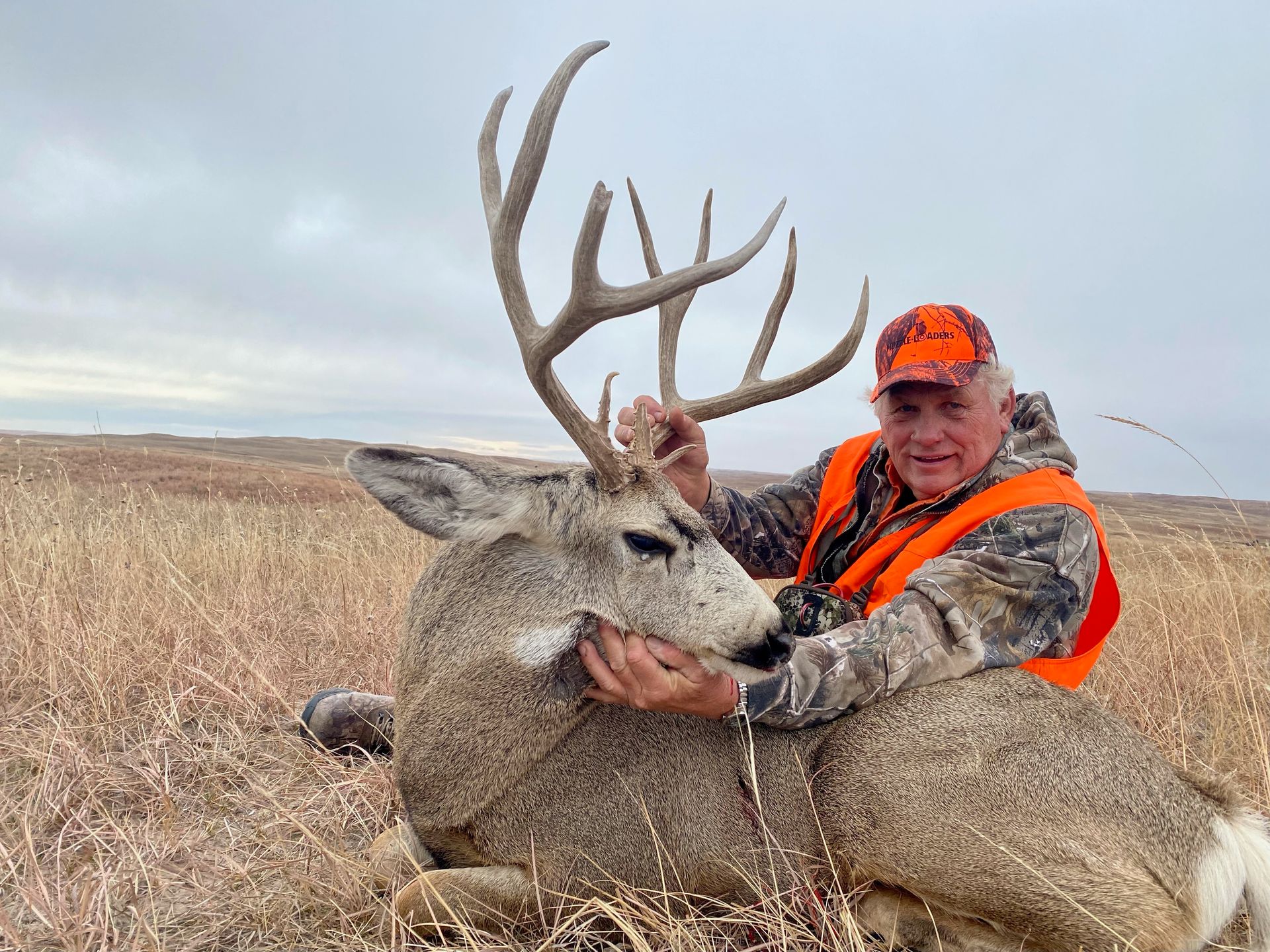 Man in orange vest smiles, holding antlers of a large dead deer in a grassy field under a cloudy sky.
