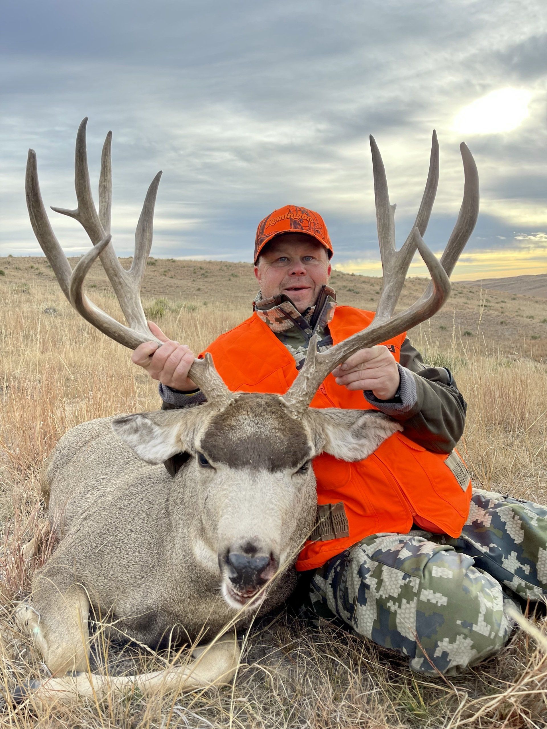 Hunter in orange vest smiles, holding large mule deer antlers in a grassy field.
