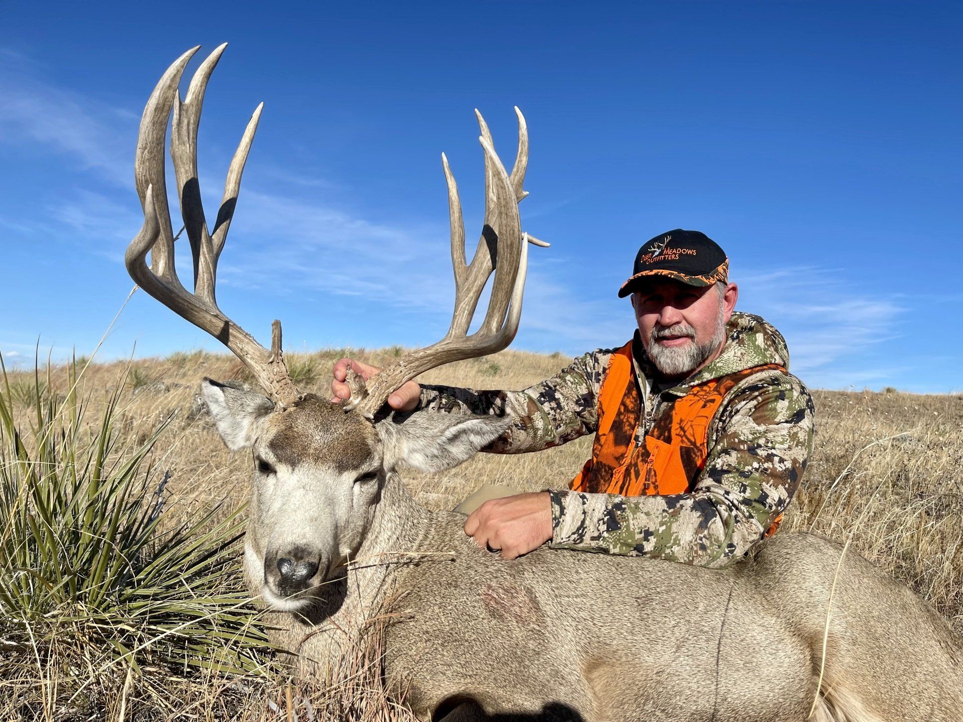 Man in hunting gear kneels next to a large buck he has harvested.