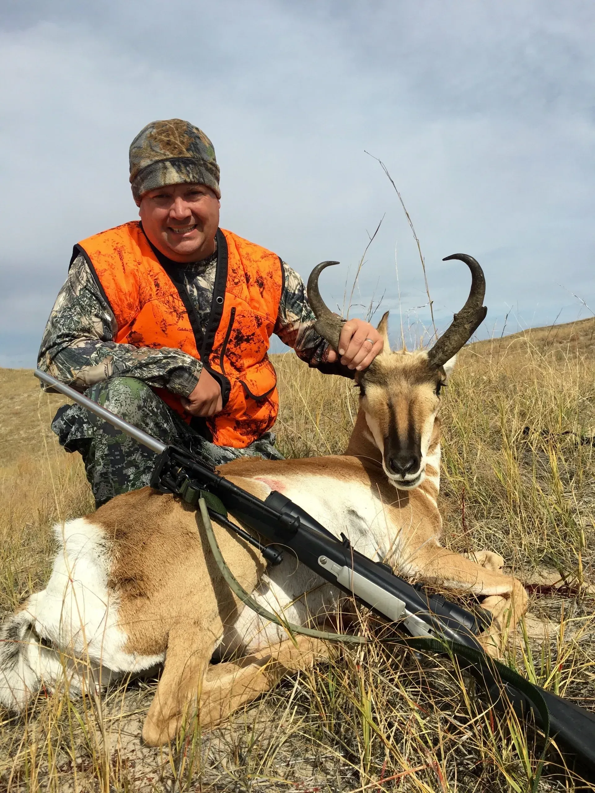 Hunter in camouflage and orange vest with a harvested pronghorn antelope and rifle on grassy terrain.