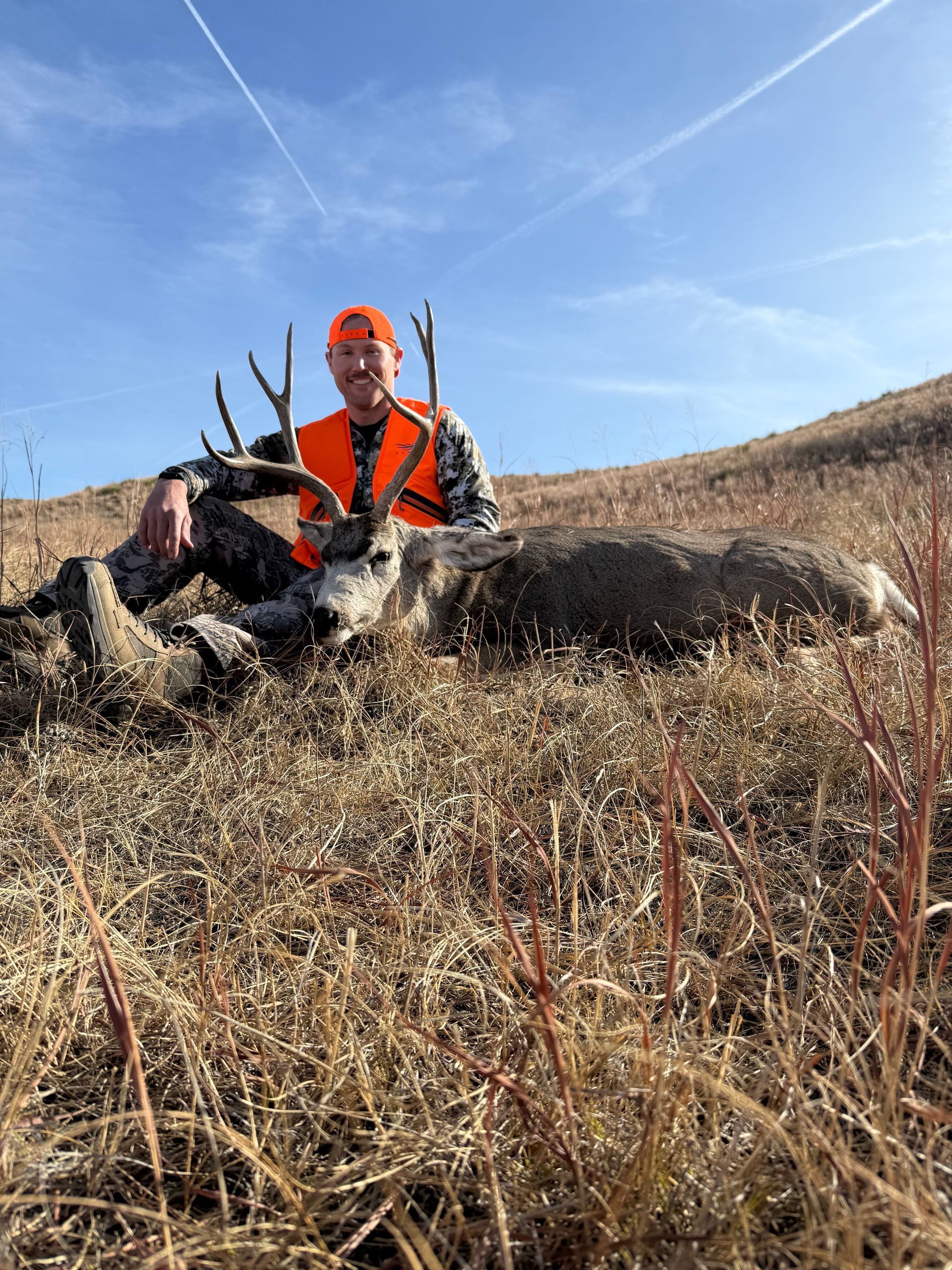Hunter in orange vest smiles next to a deer on a grassy hillside under a blue sky.