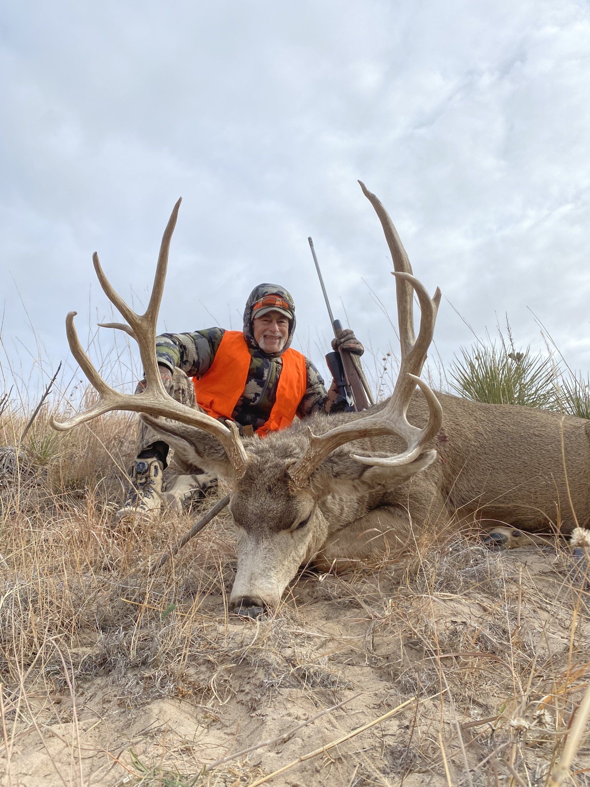 Hunter in orange vest, smiling, poses with large buck, rifle in hand, on dry, grassy terrain.