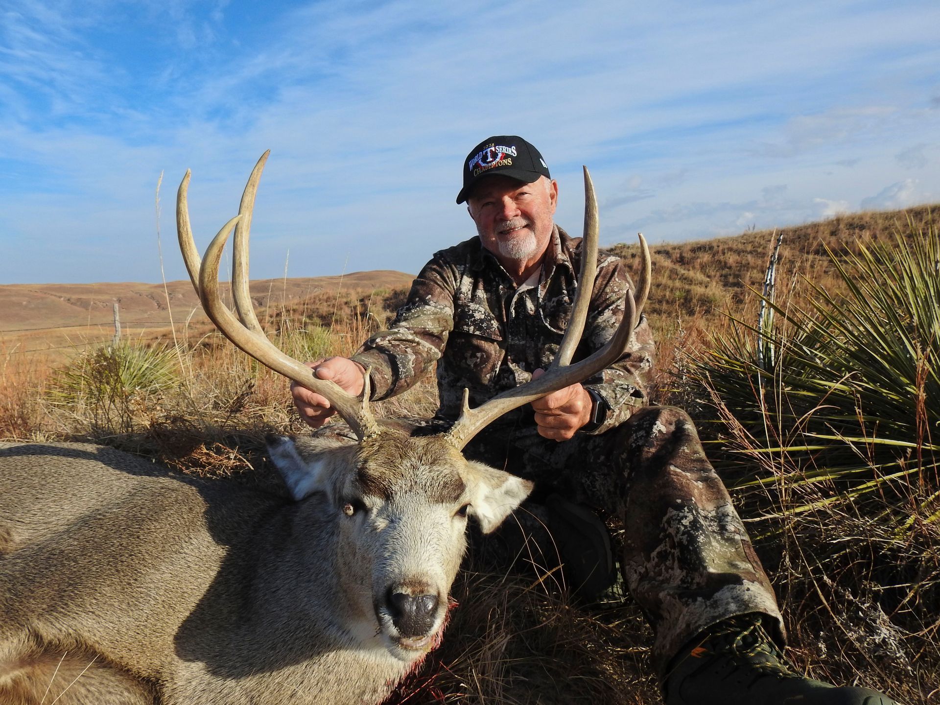 Hunter poses with a large buck; outdoors on a sunny day.