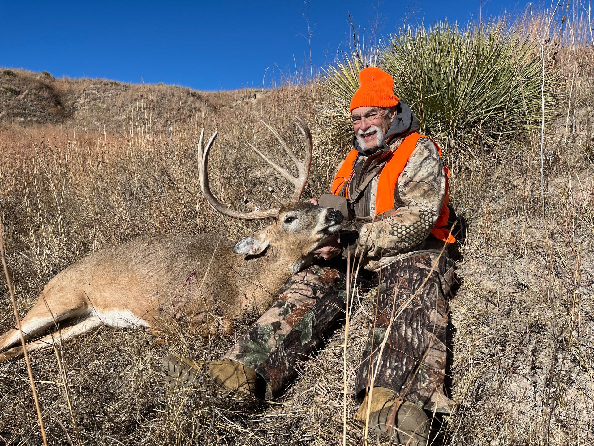 Hunter in orange vest and hat with a large buck in grassy field.