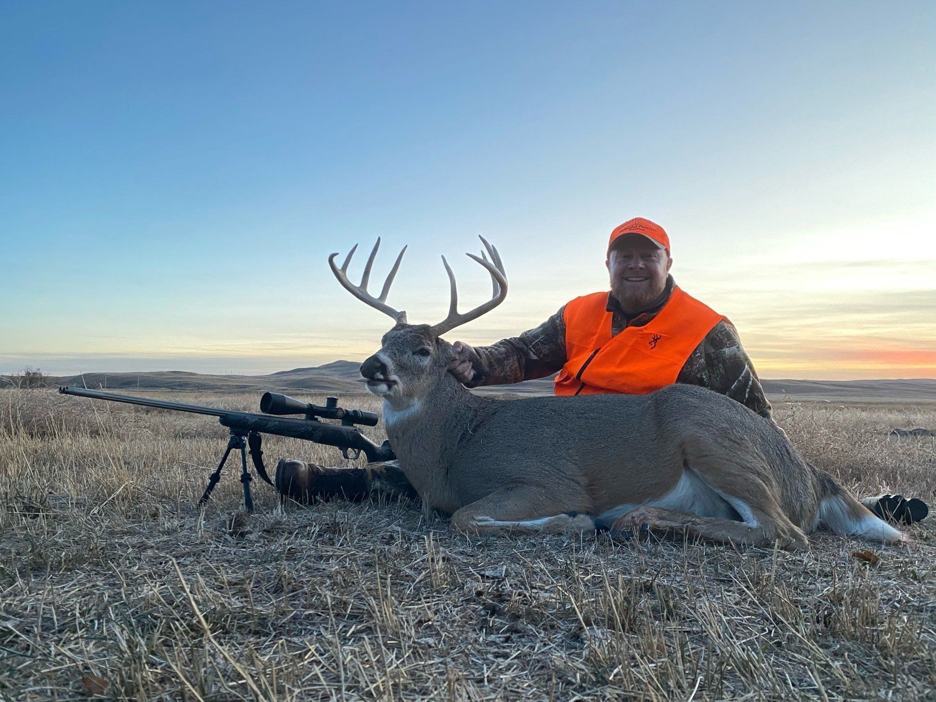 Hunter in orange vest kneels next to large buck with impressive antlers in a field at sunset, rifle nearby.