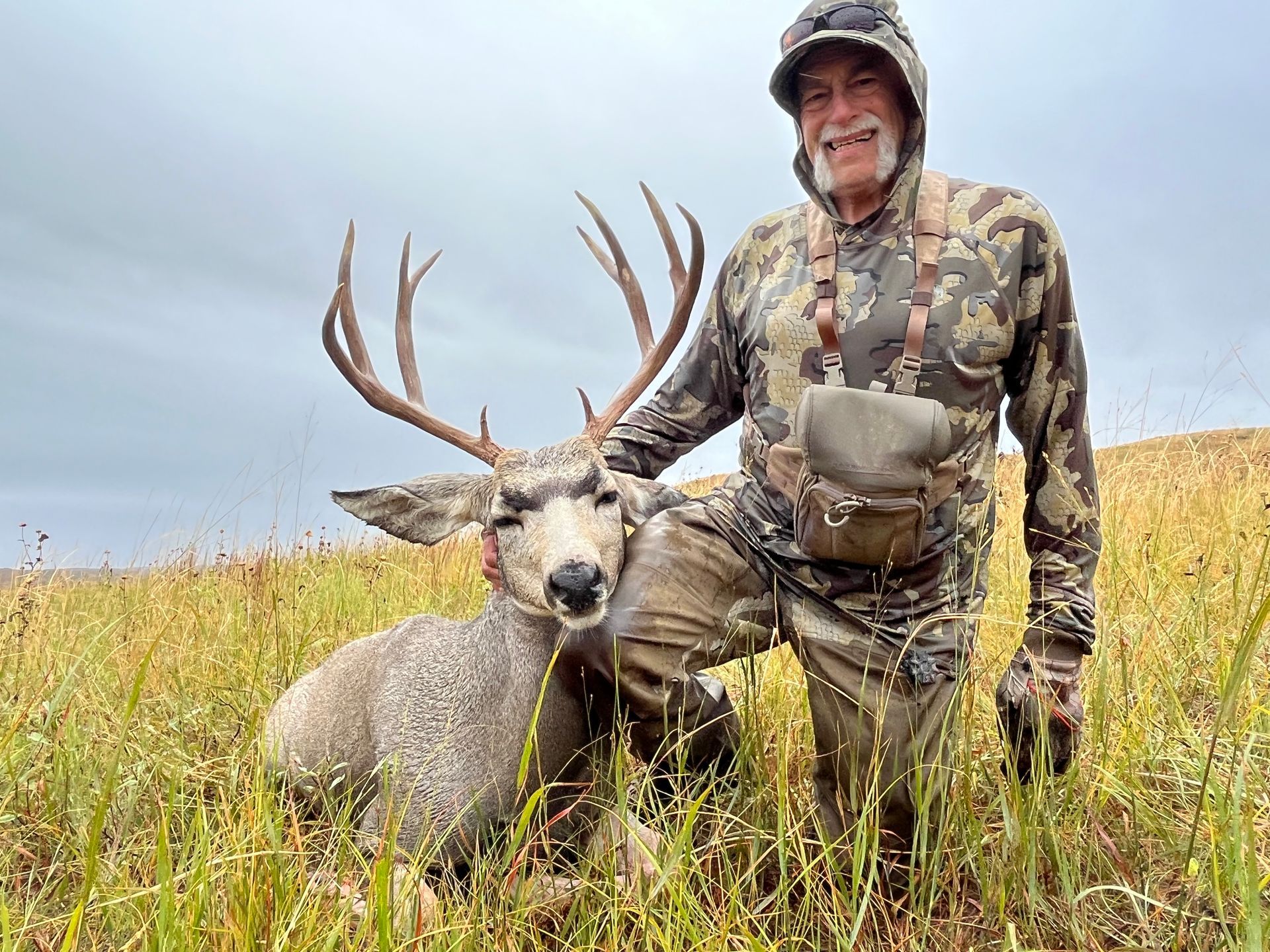Hunter kneels beside a large buck in tall grass. The man wears camo clothing and smiles. Cloudy, outdoor setting.