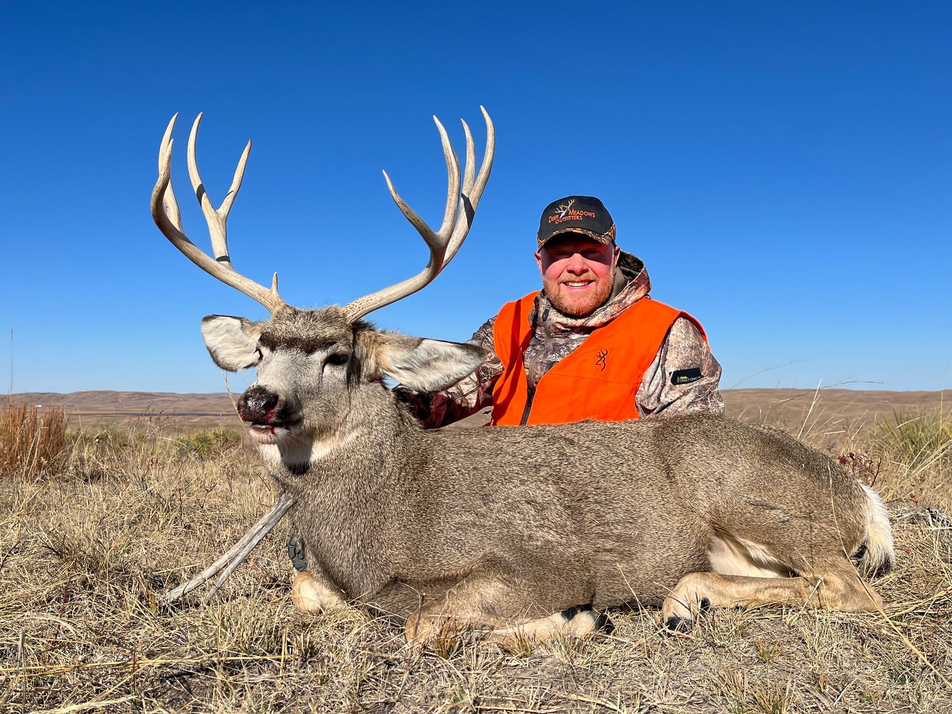 Man in orange vest smiles next to a large dead deer with impressive antlers in a field.