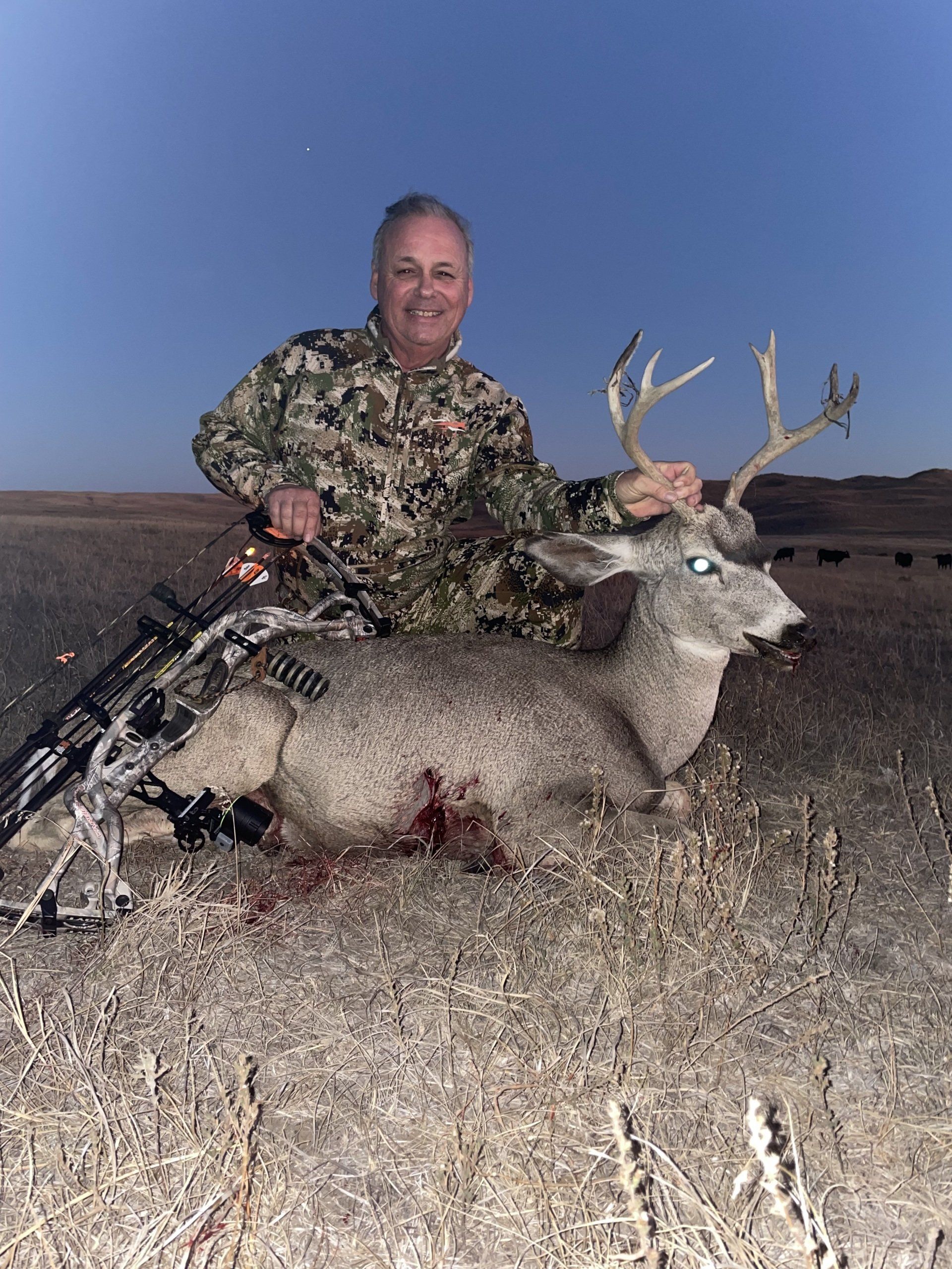 Man in camo smiling, kneeling beside a harvested deer with antlers, holding a bow, against a night sky.