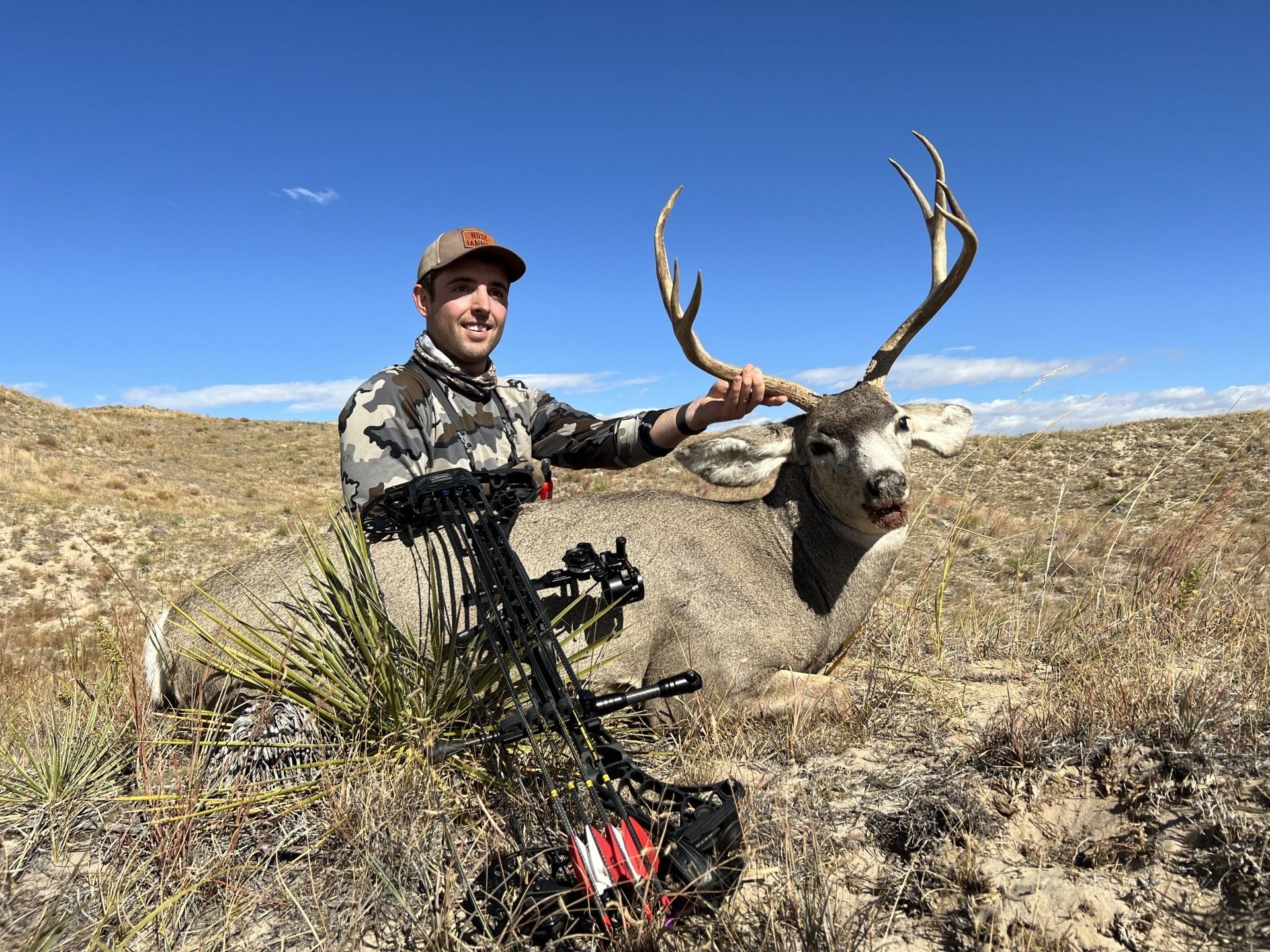 Hunter with a large deer he harvested in a sunny, arid landscape. He smiles while holding the antlers.