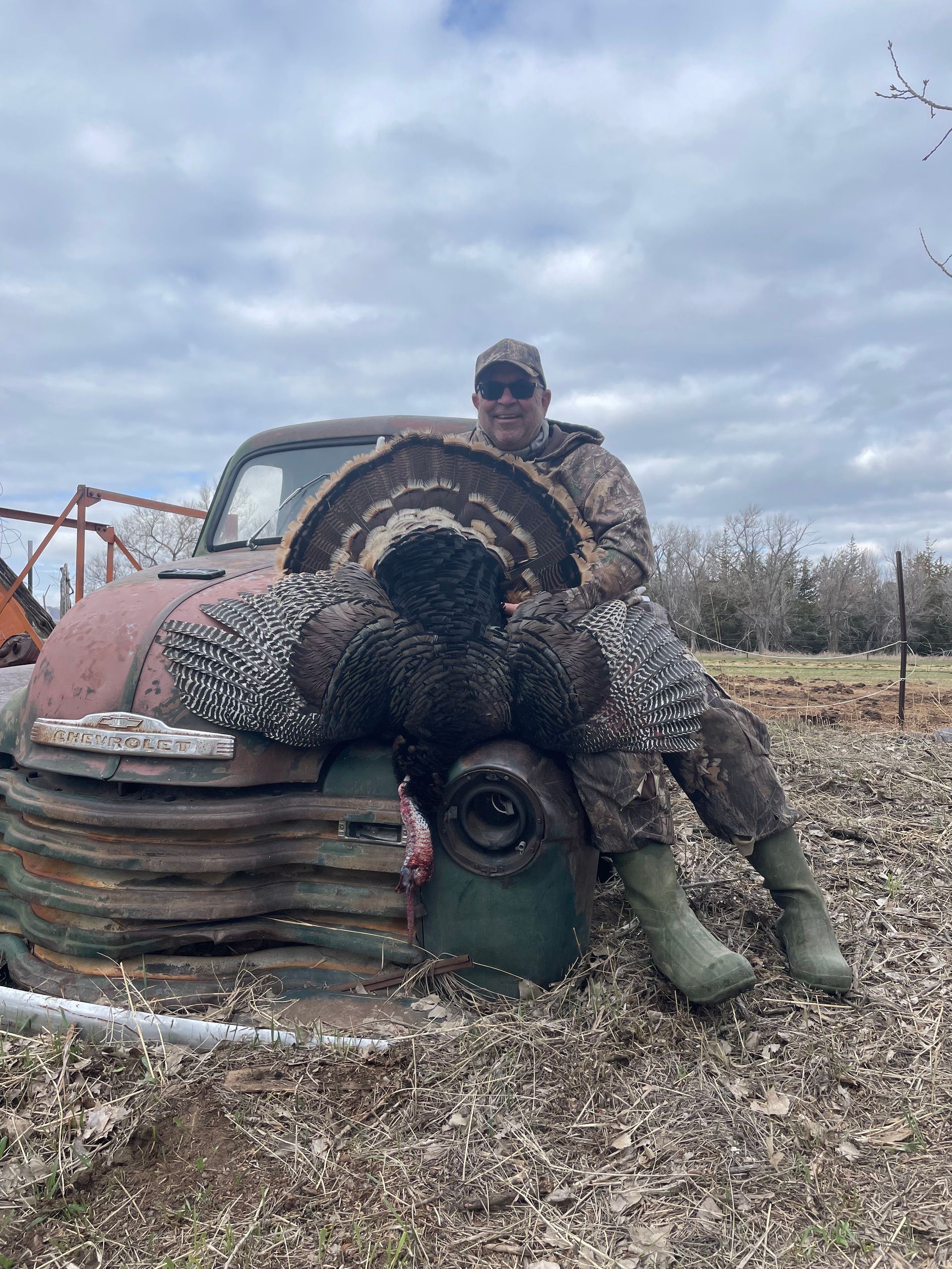 Man in camo holds a large turkey in front of an old truck in a field.