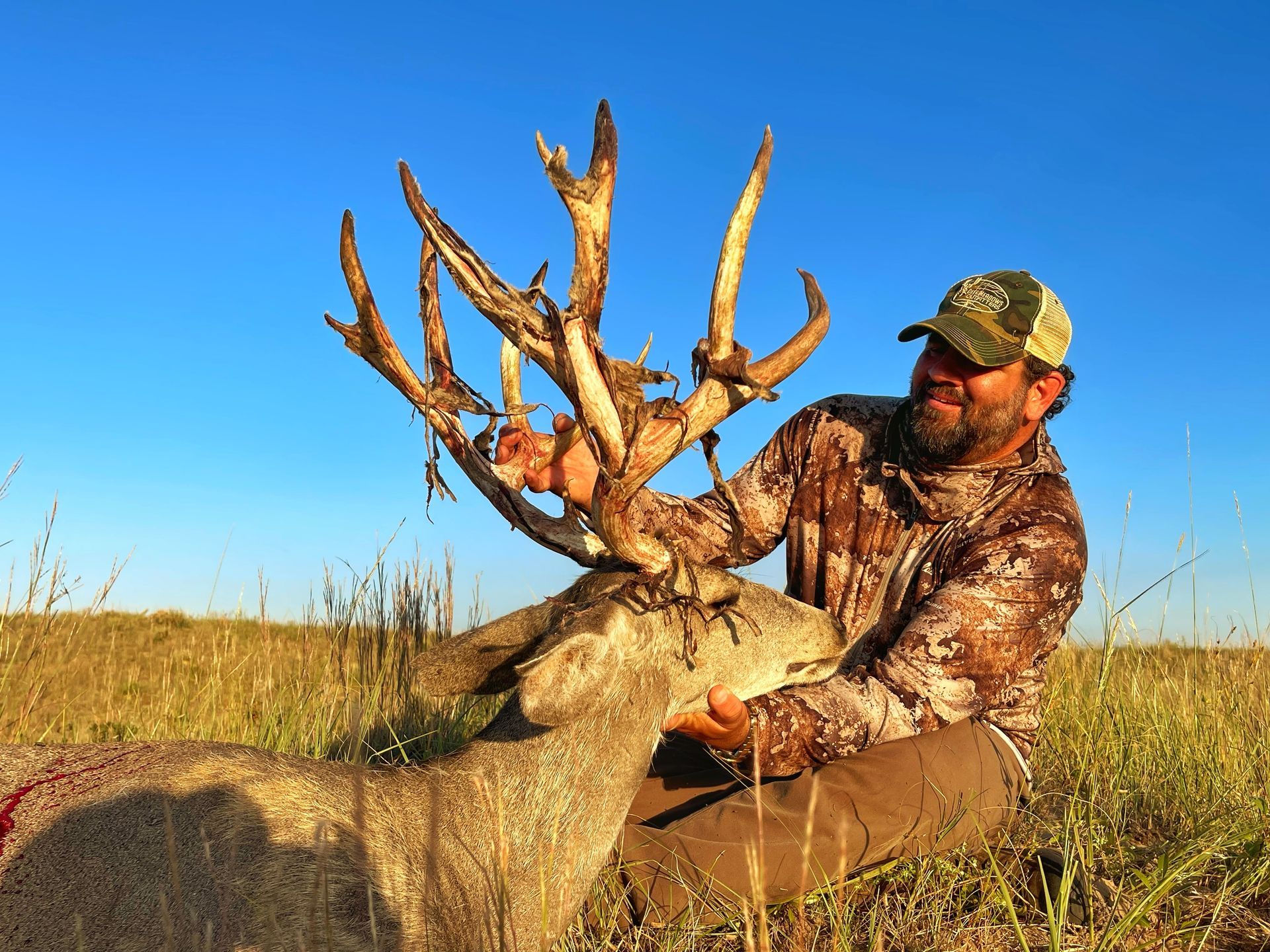 Man kneeling beside a large mule deer with impressive antlers, smiling, in a field under a blue sky.