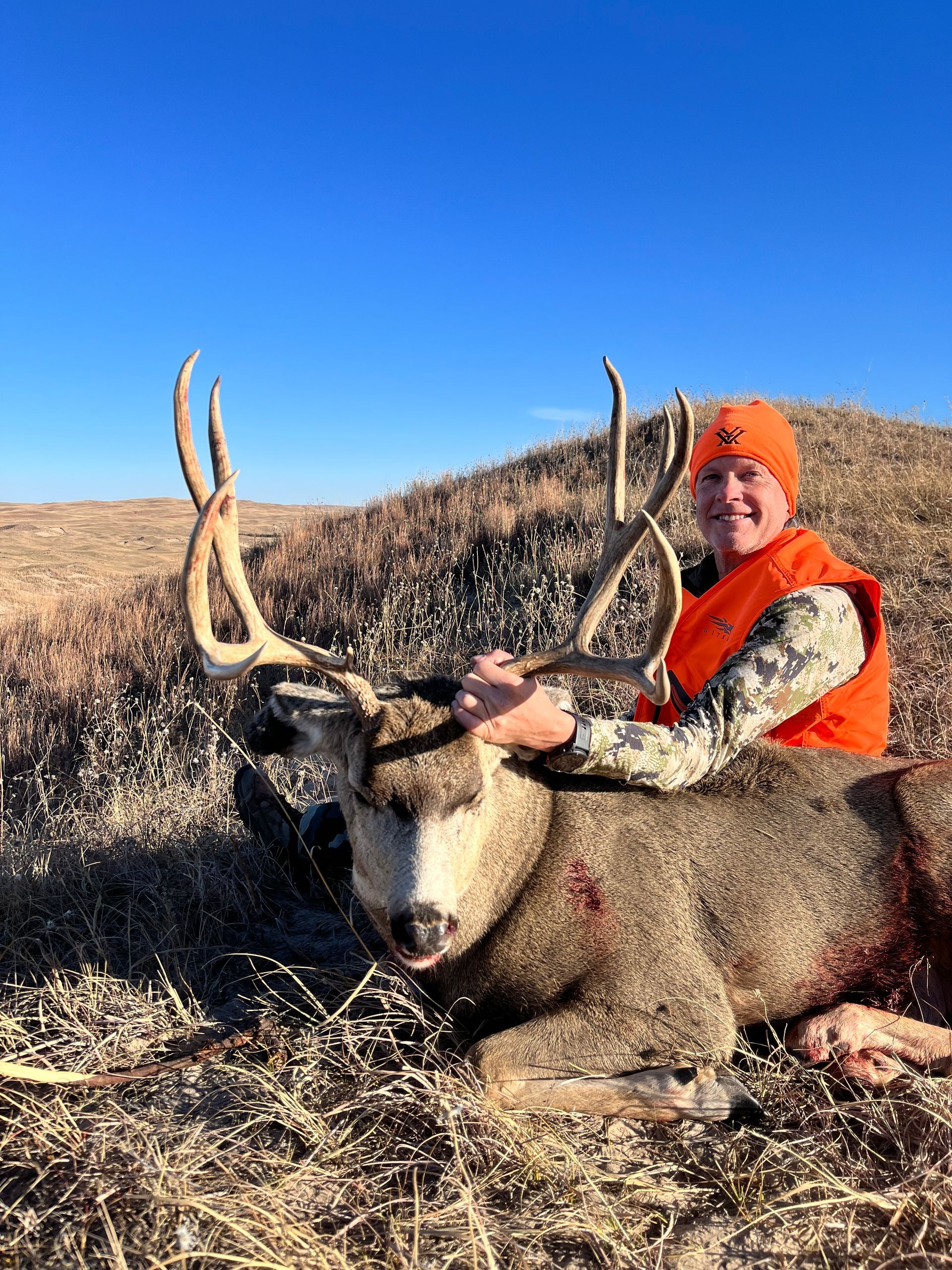 Hunter in orange vest smiles next to a large deer with impressive antlers. Blue sky. Brown field.