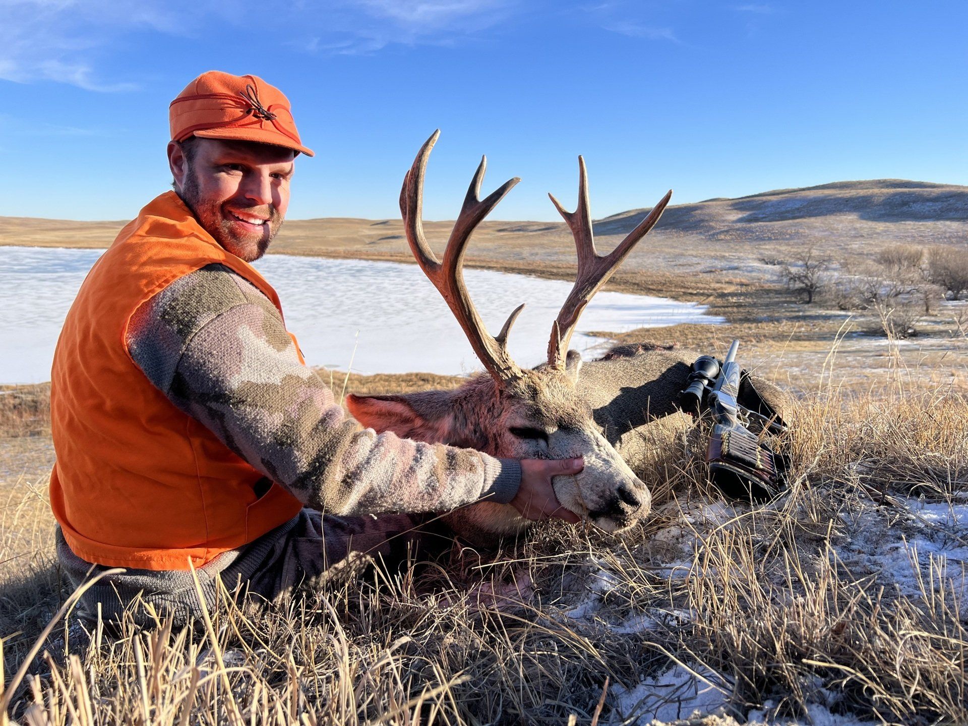 Hunter kneels beside a harvested buck with large antlers, orange vest and cap, snowy landscape.