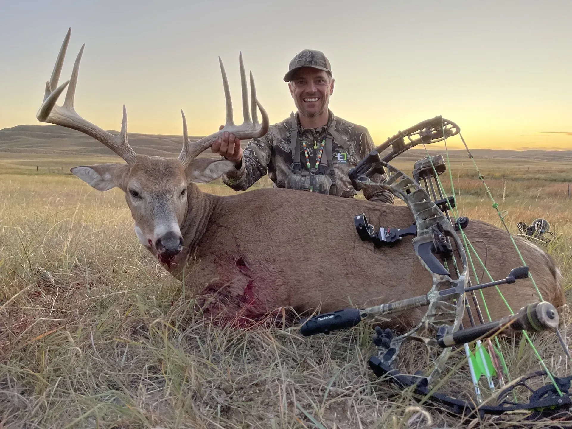 Man smiles, holding large buck's antlers. Shot deer lies in grassy field with bow. Sunset.
