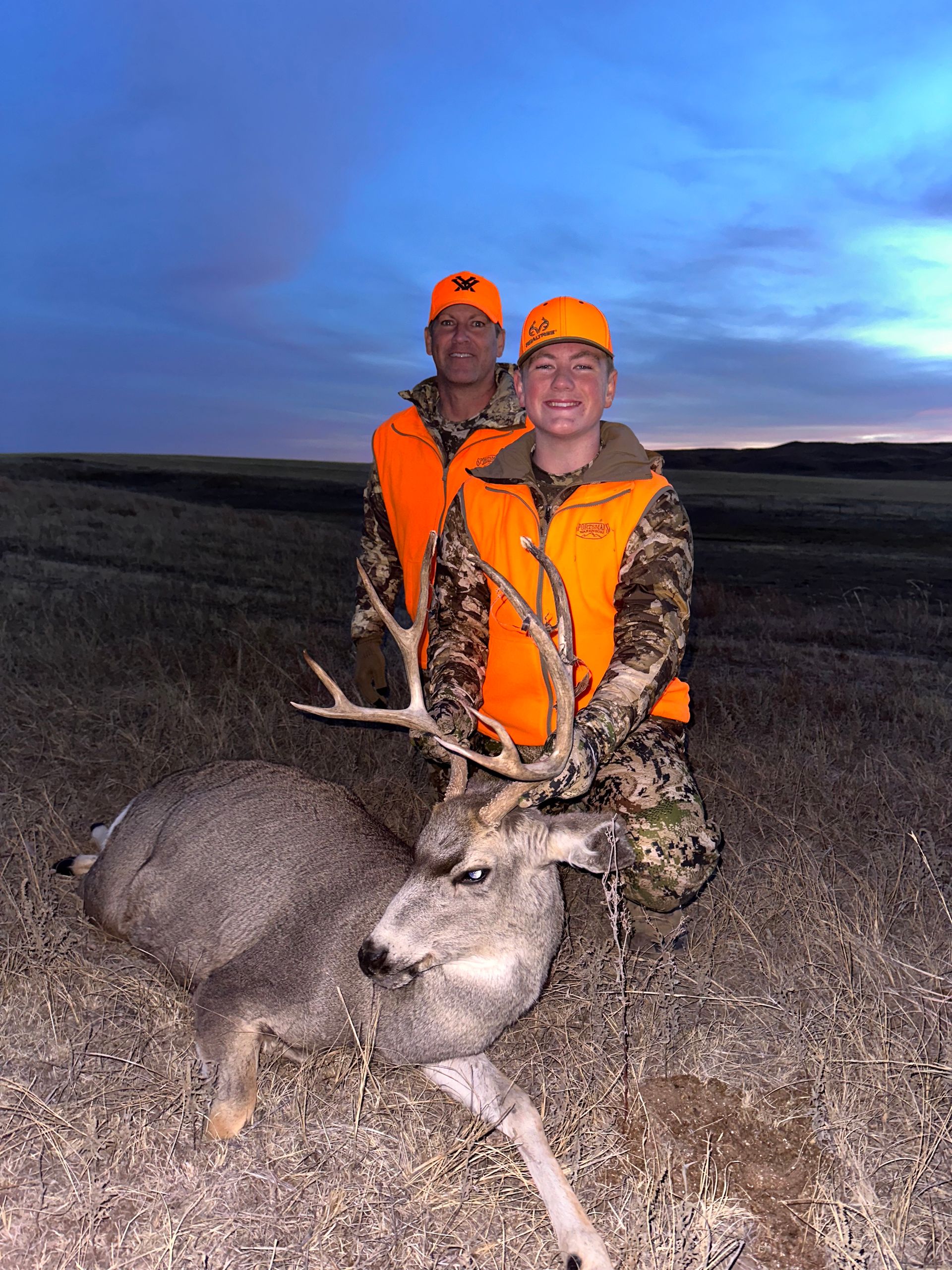 Two hunters in orange vests pose with a harvested deer at dusk.
