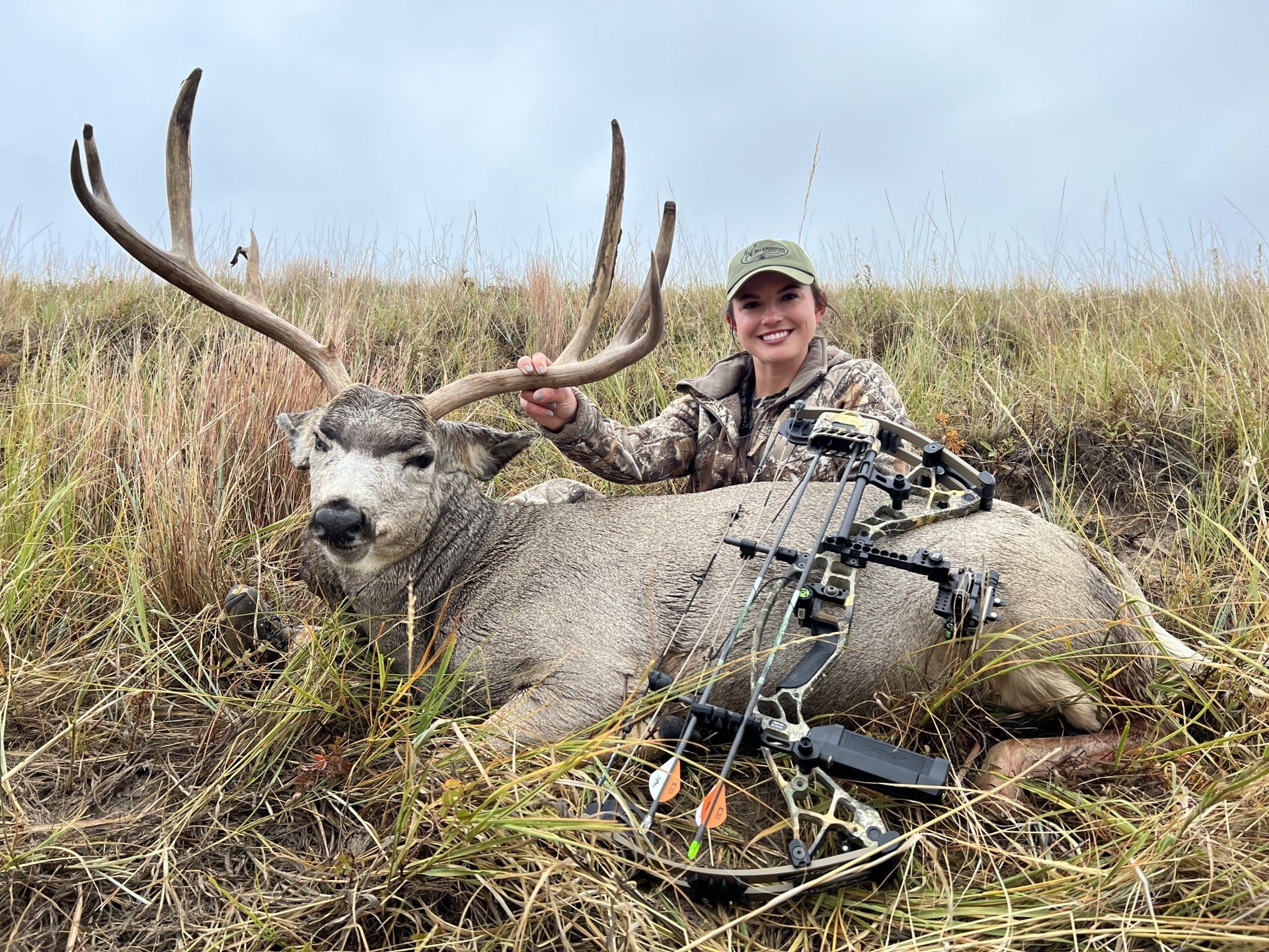 Woman in camo smiles, holding large deer antlers, next to a fallen deer and bow in grassy field.