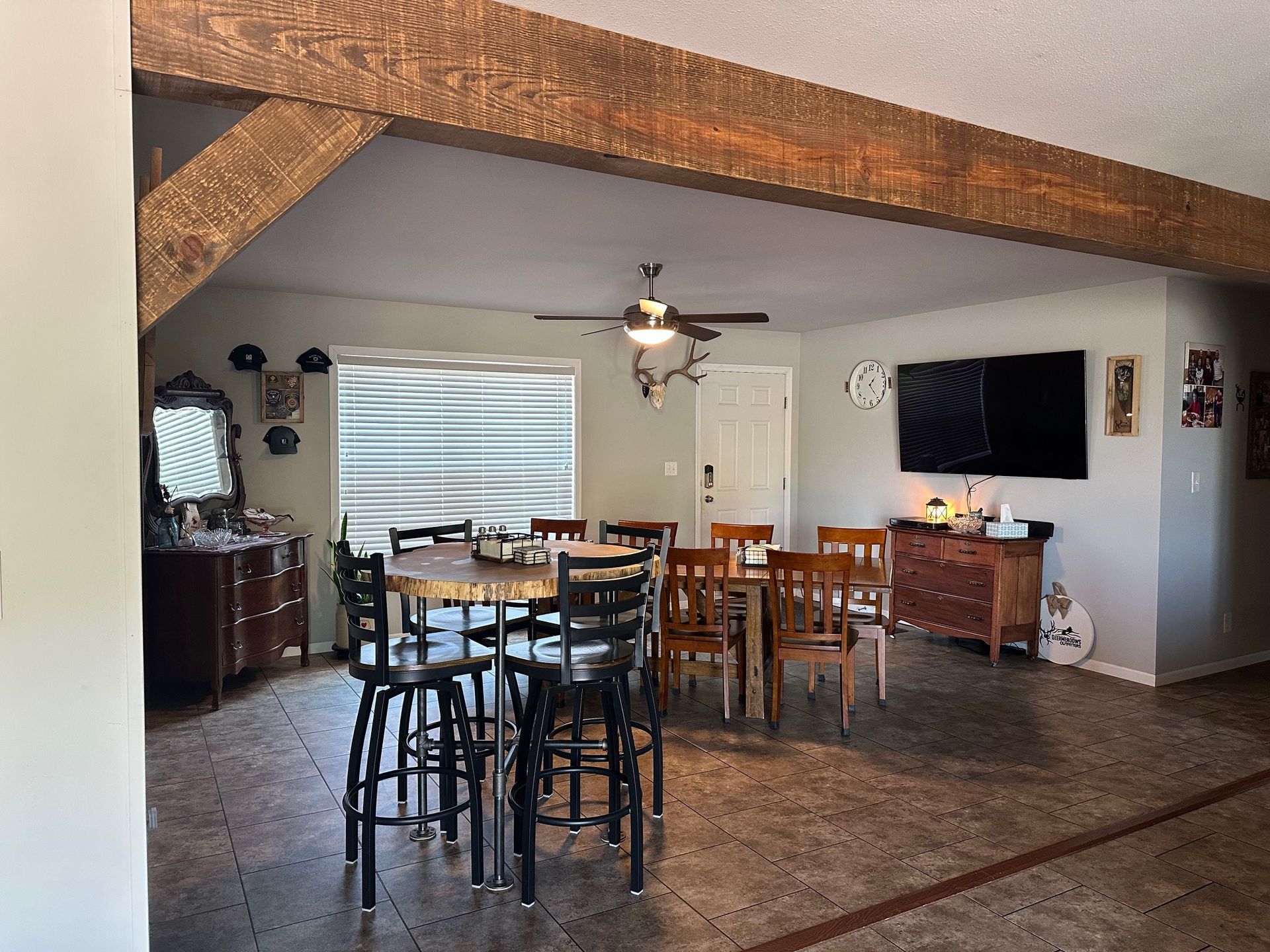 A dining area with a round table, chairs, and a TV on the wall. Dark brown wooden beams overhead.