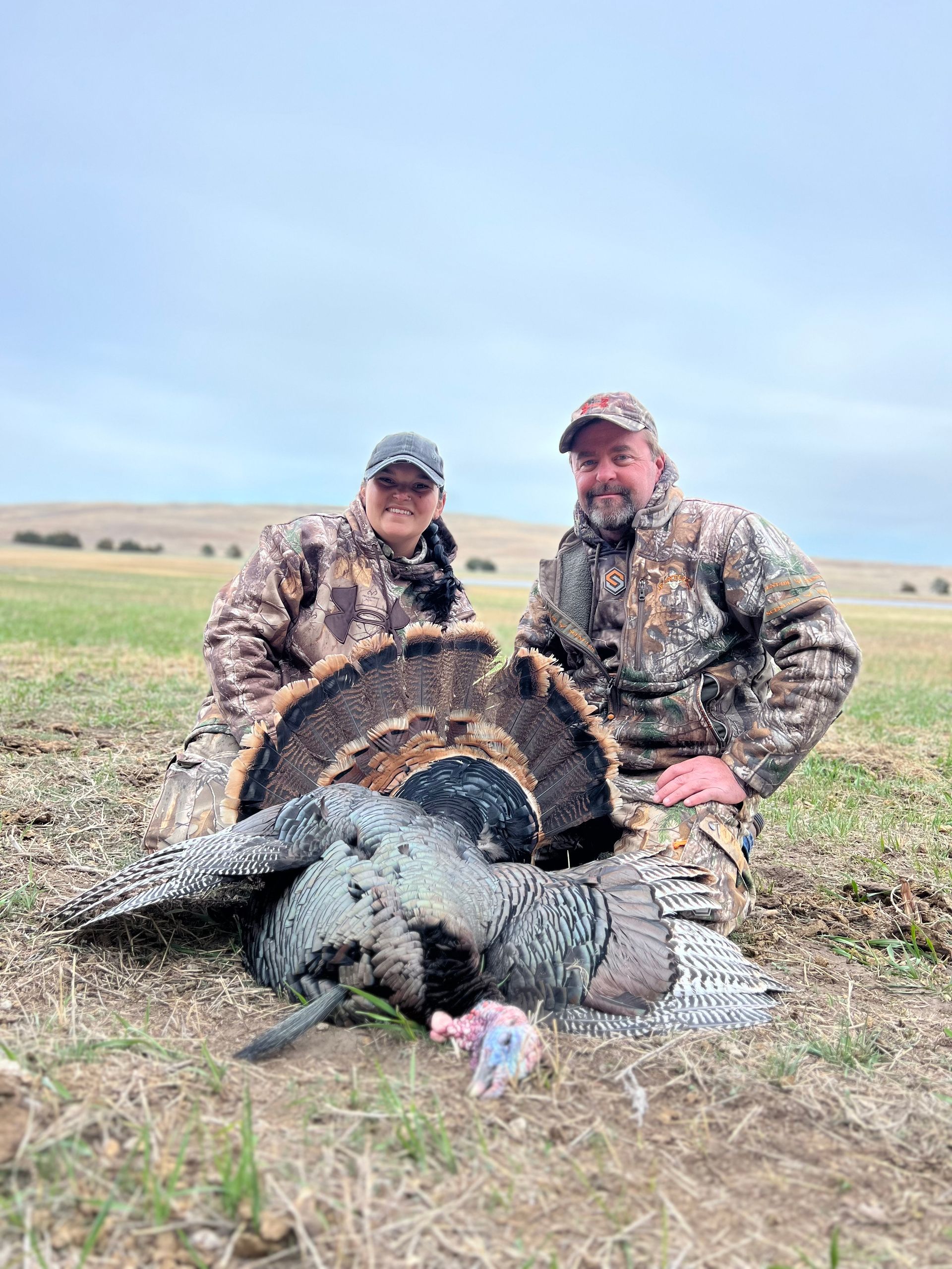 Two people in camo kneel next to a harvested turkey in a grassy field under a cloudy sky.