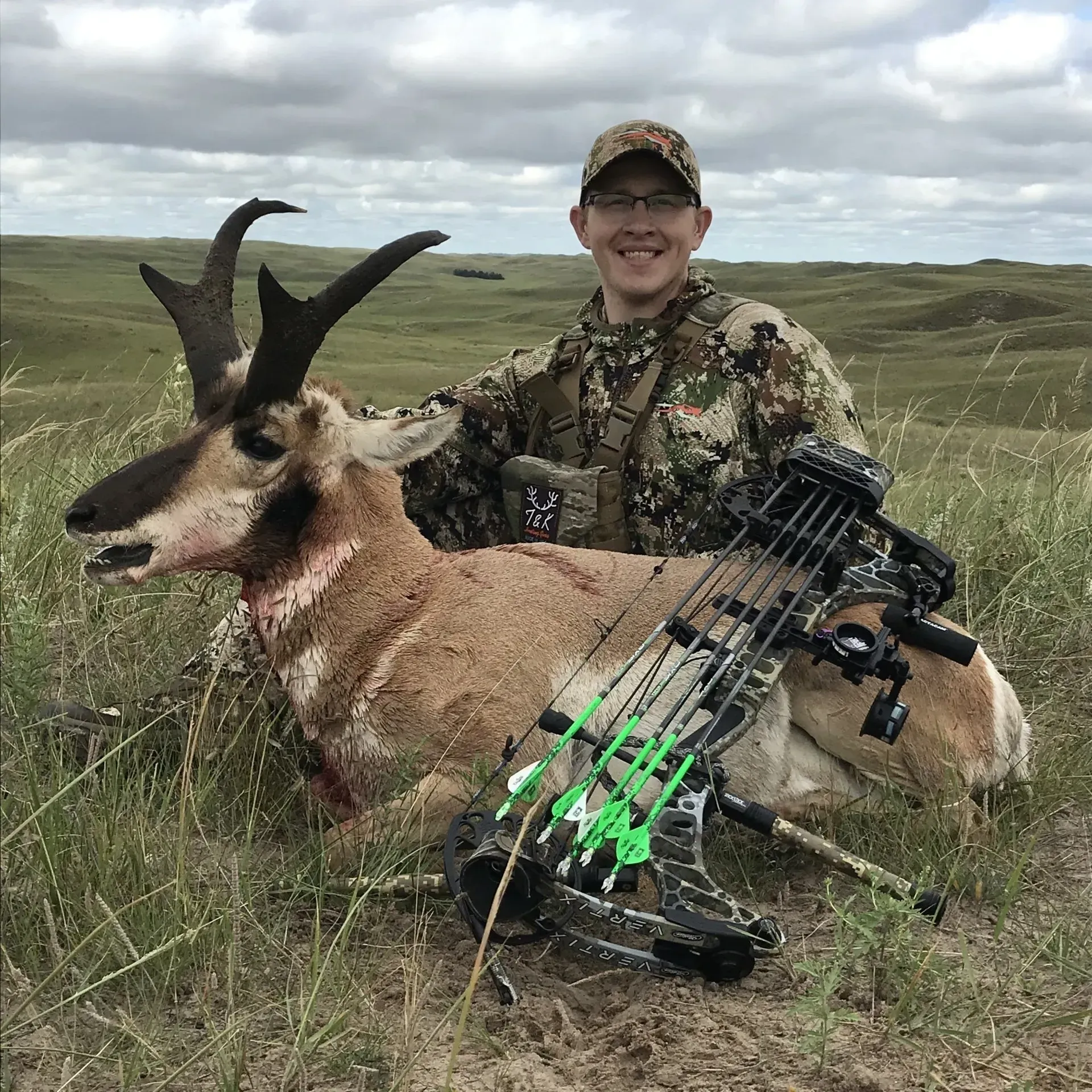 Man in camo with a pronghorn antelope he shot with a bow in grassy field. He is smiling.