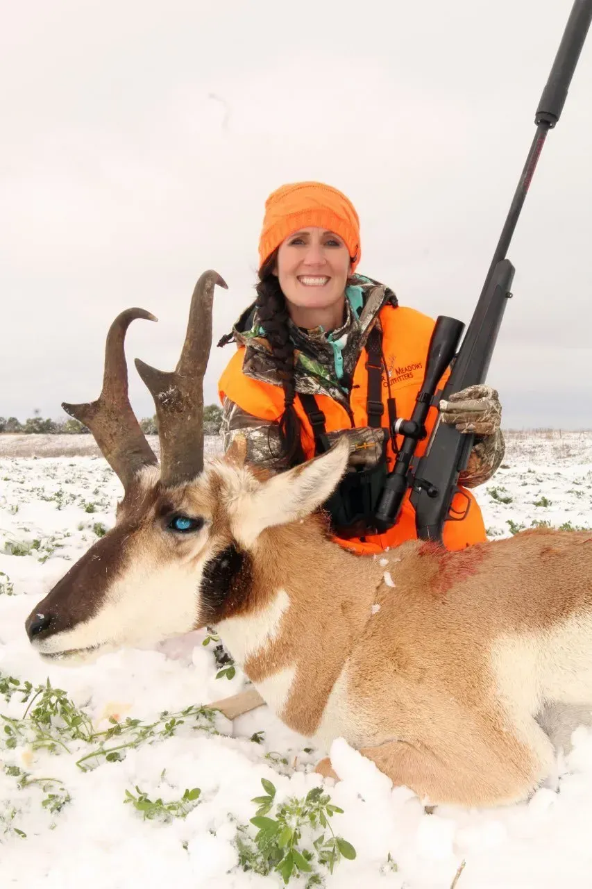 Woman in orange hunting gear smiles, posing with an antelope she harvested on a snowy field.
