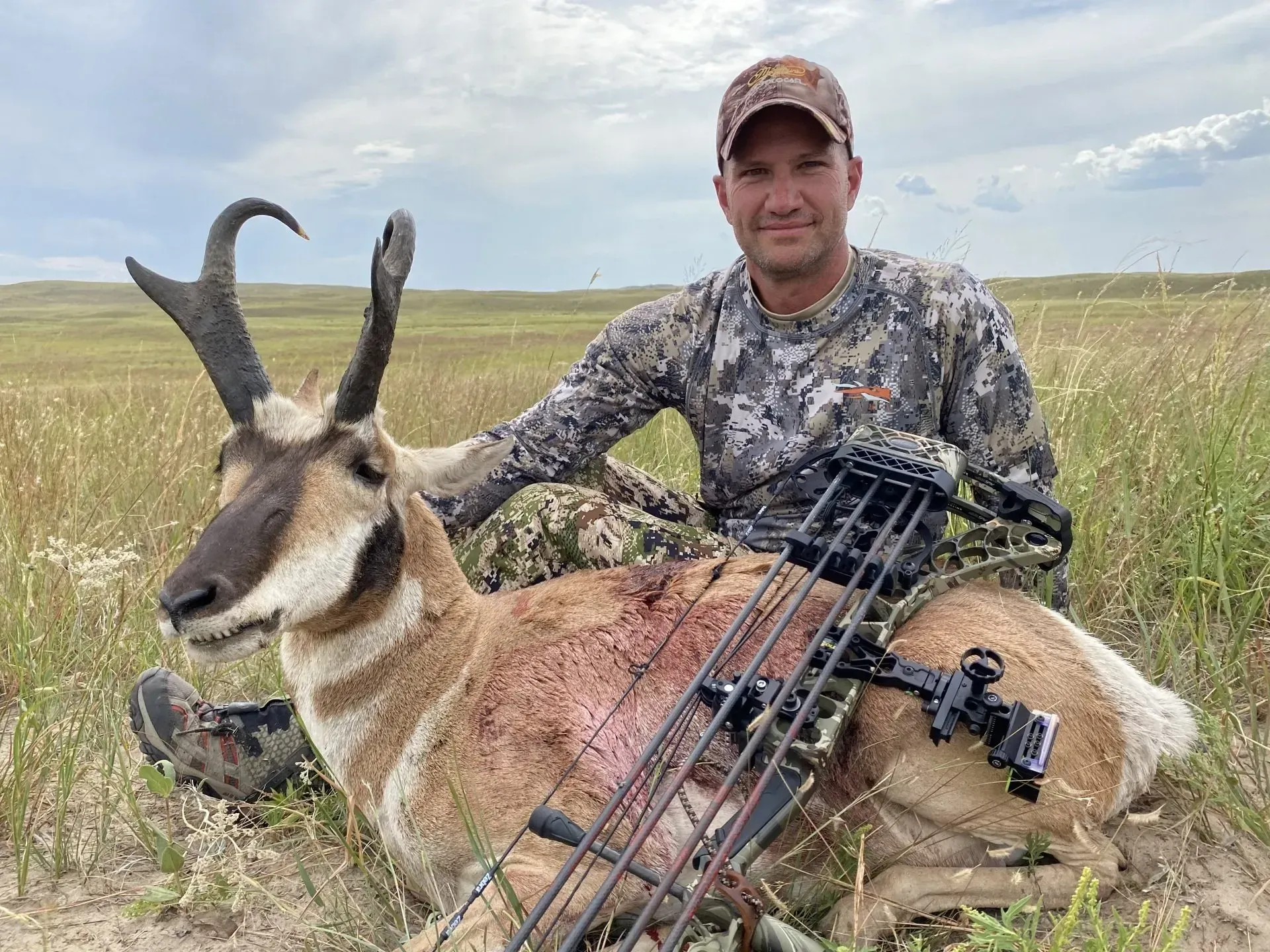 Hunter with a bow, posing with a harvested pronghorn antelope in a grassy field.