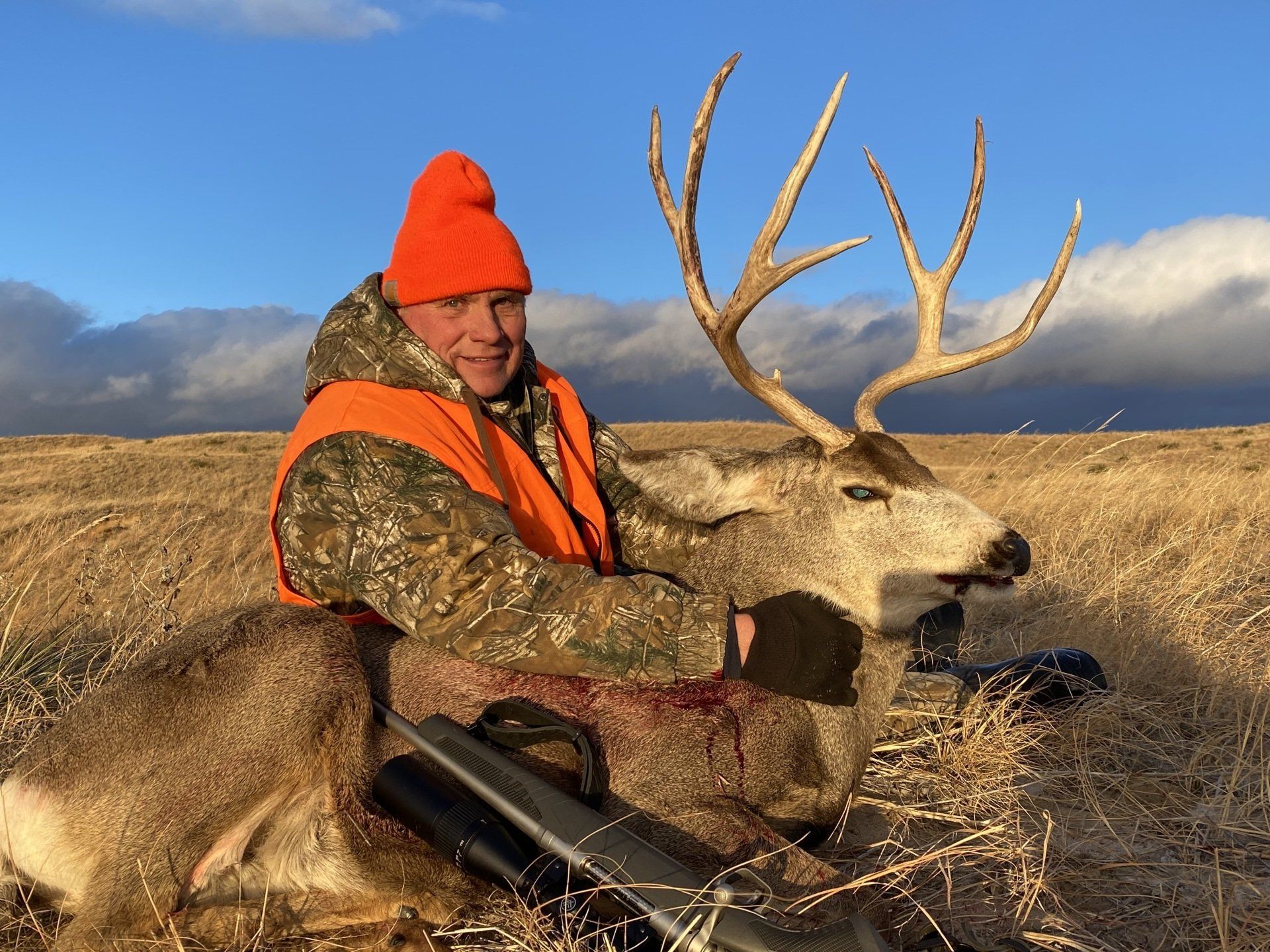 Hunter in orange vest kneels beside a large deer with impressive antlers on a grassy plain.