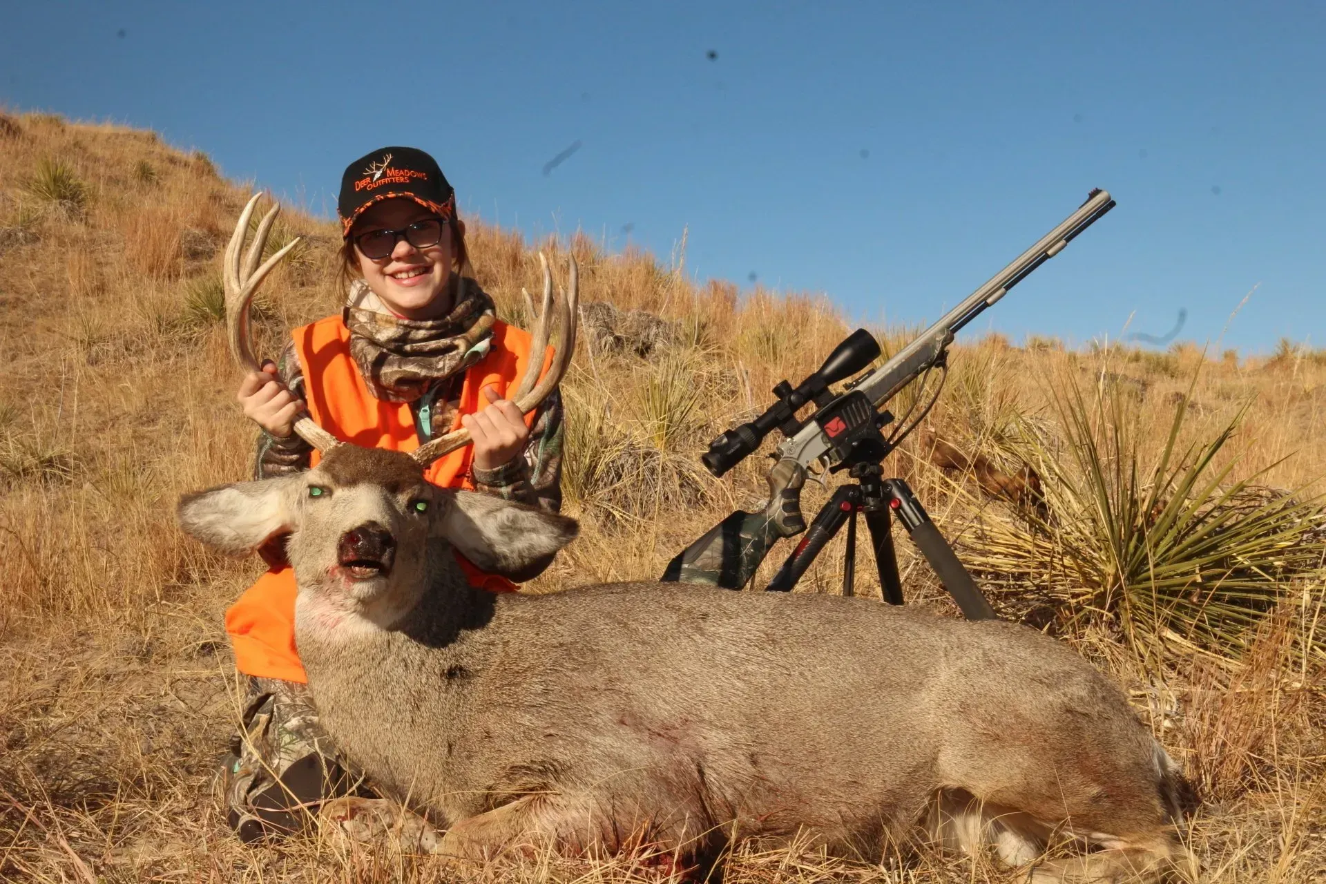 Young hunter kneels with a deer, holding its antlers. Rifle on tripod in a grassy field, wearing orange vest.