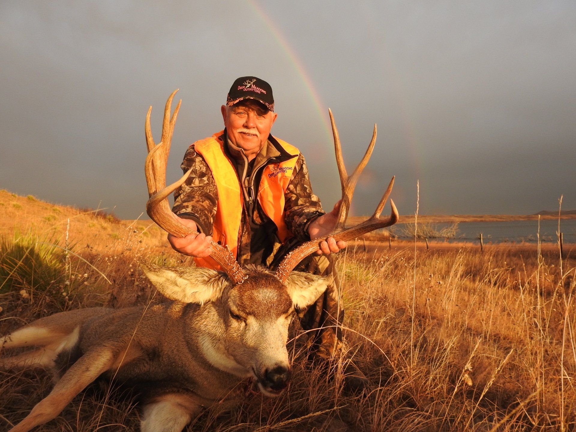 Man in hunting gear with a large buck under a rainbow.  Open field with a lake in background.