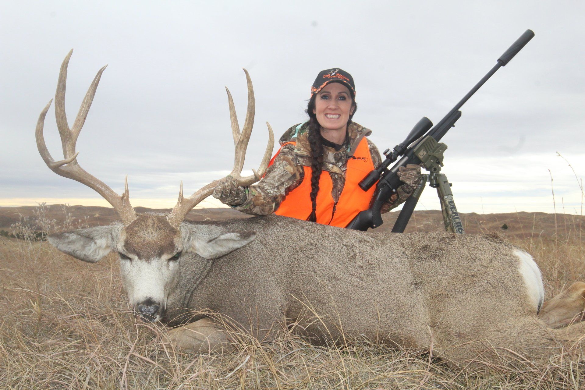 Woman in orange vest with a deer, rifle in setting of tan tall grass.