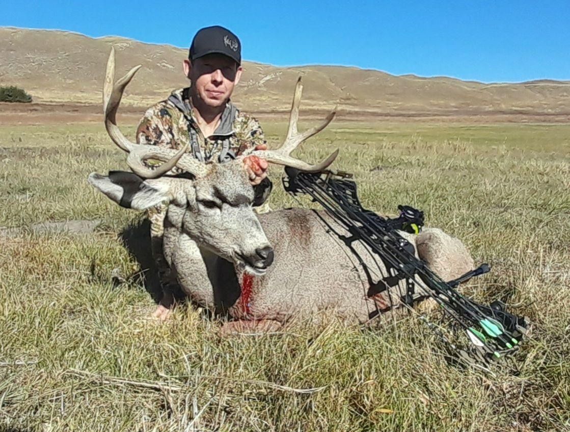 Hunter posing with a harvested mule deer buck and bow in a grassy field, sunny day.