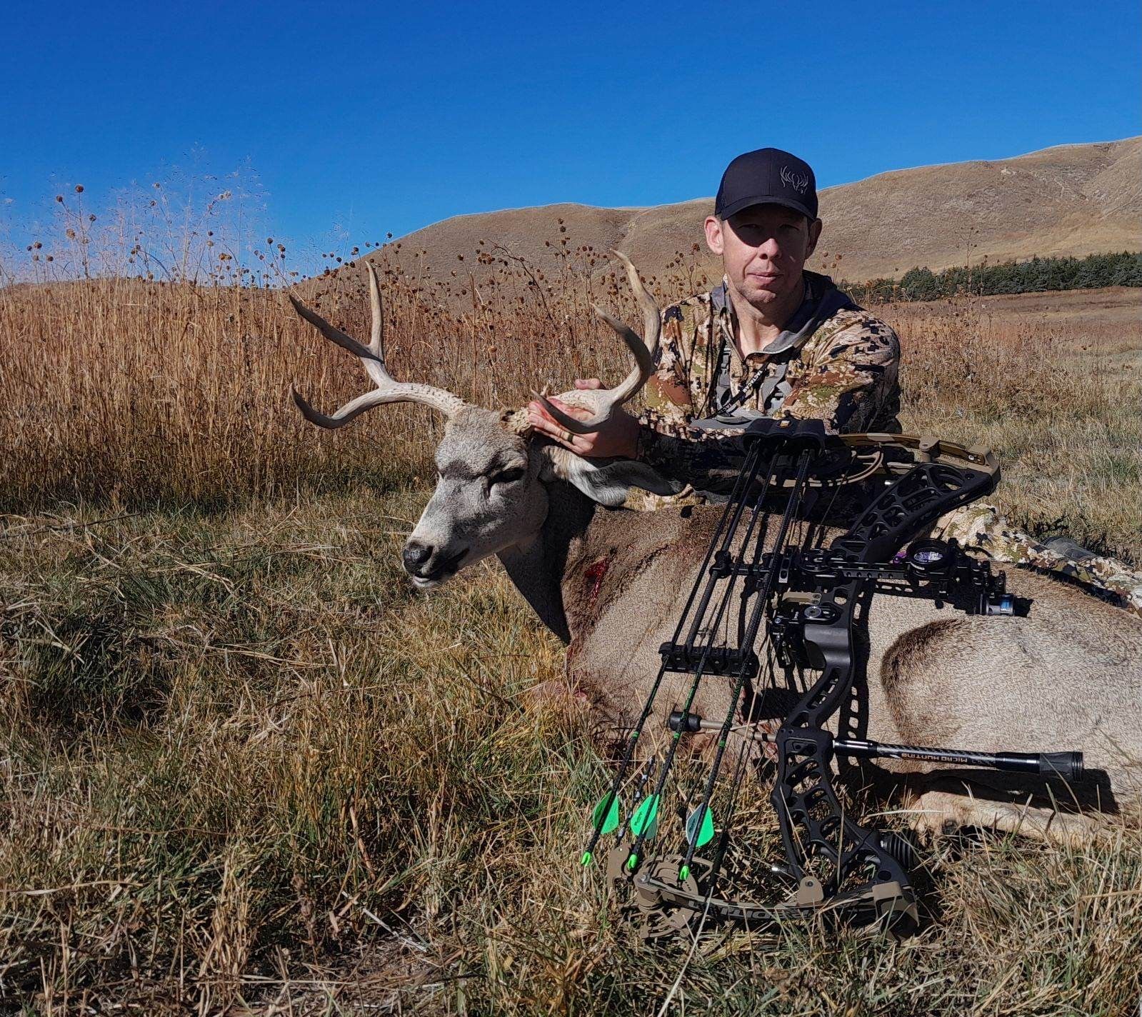 Hunter with a harvested deer in a field. He holds its antlers, and his bow is in the foreground.