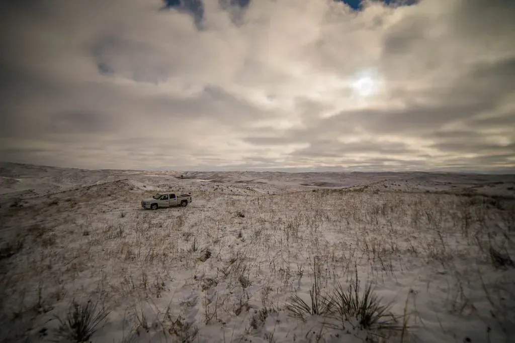 A snowy field under a cloudy sky, with a white pickup truck driving through it.
