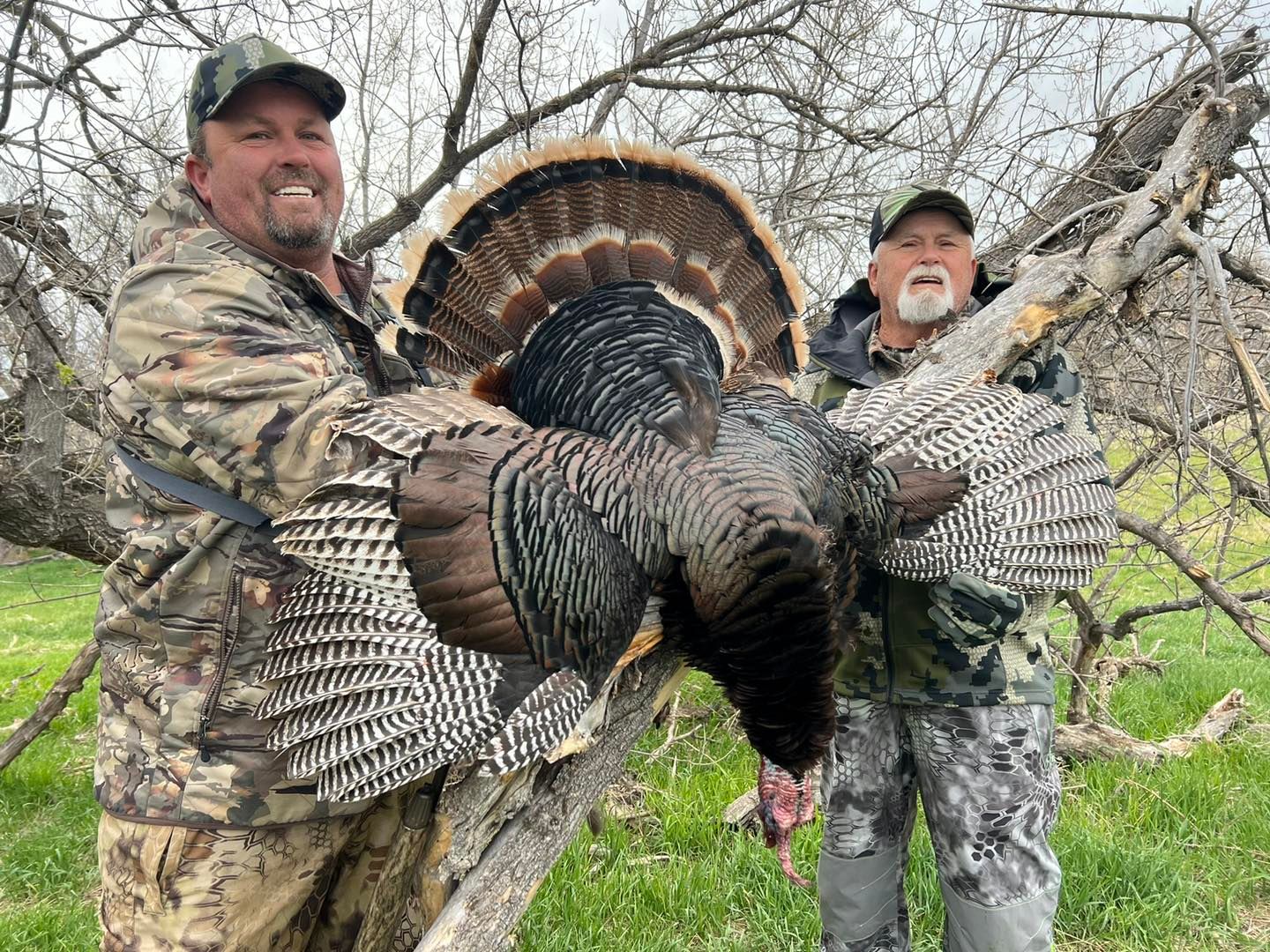 Two men in camouflage hold a large wild turkey outdoors; they smile, tree branches in the background.