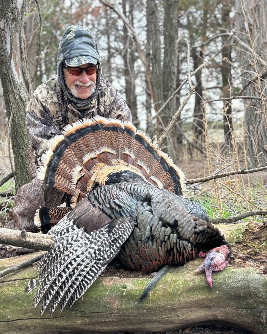 Smiling man in camo poses with a harvested wild turkey in a wooded area.