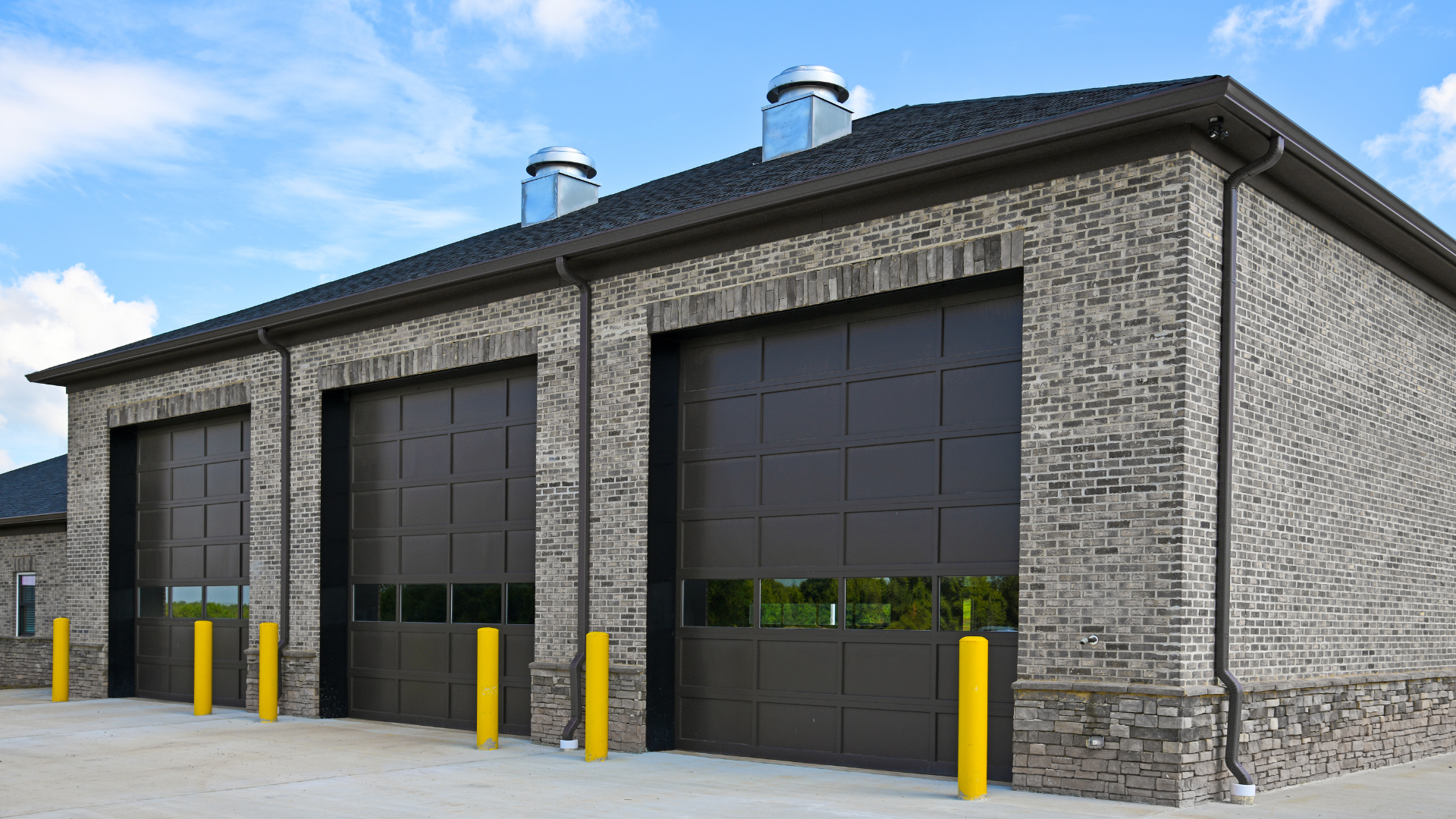 Brick industrial building with three large black garage doors and yellow bollards in front