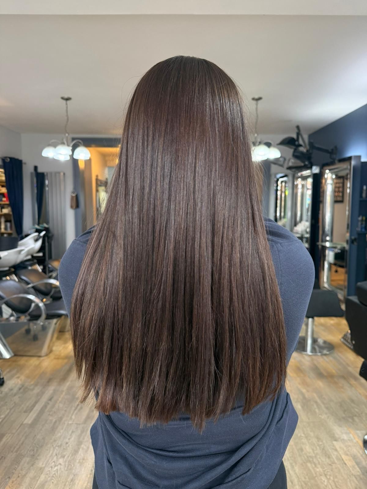 Back view of long, straight brown hair in a salon with mirrors and styling chairs in the background