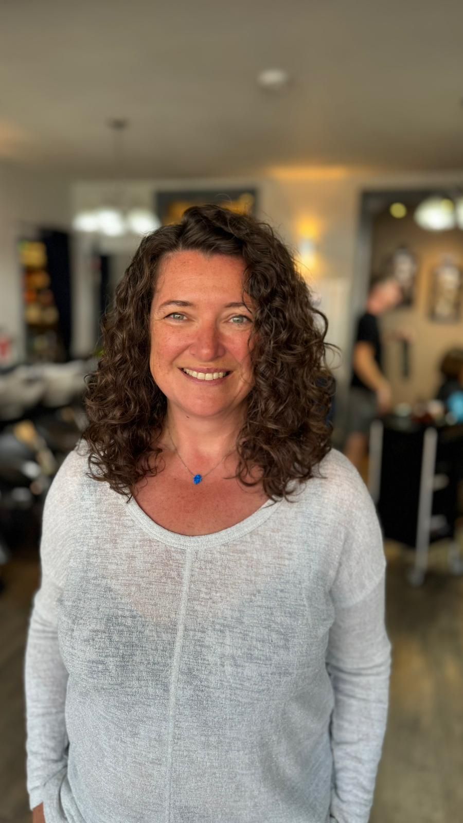 Smiling woman with curly hair in a light gray top, standing indoors in a softly lit room.