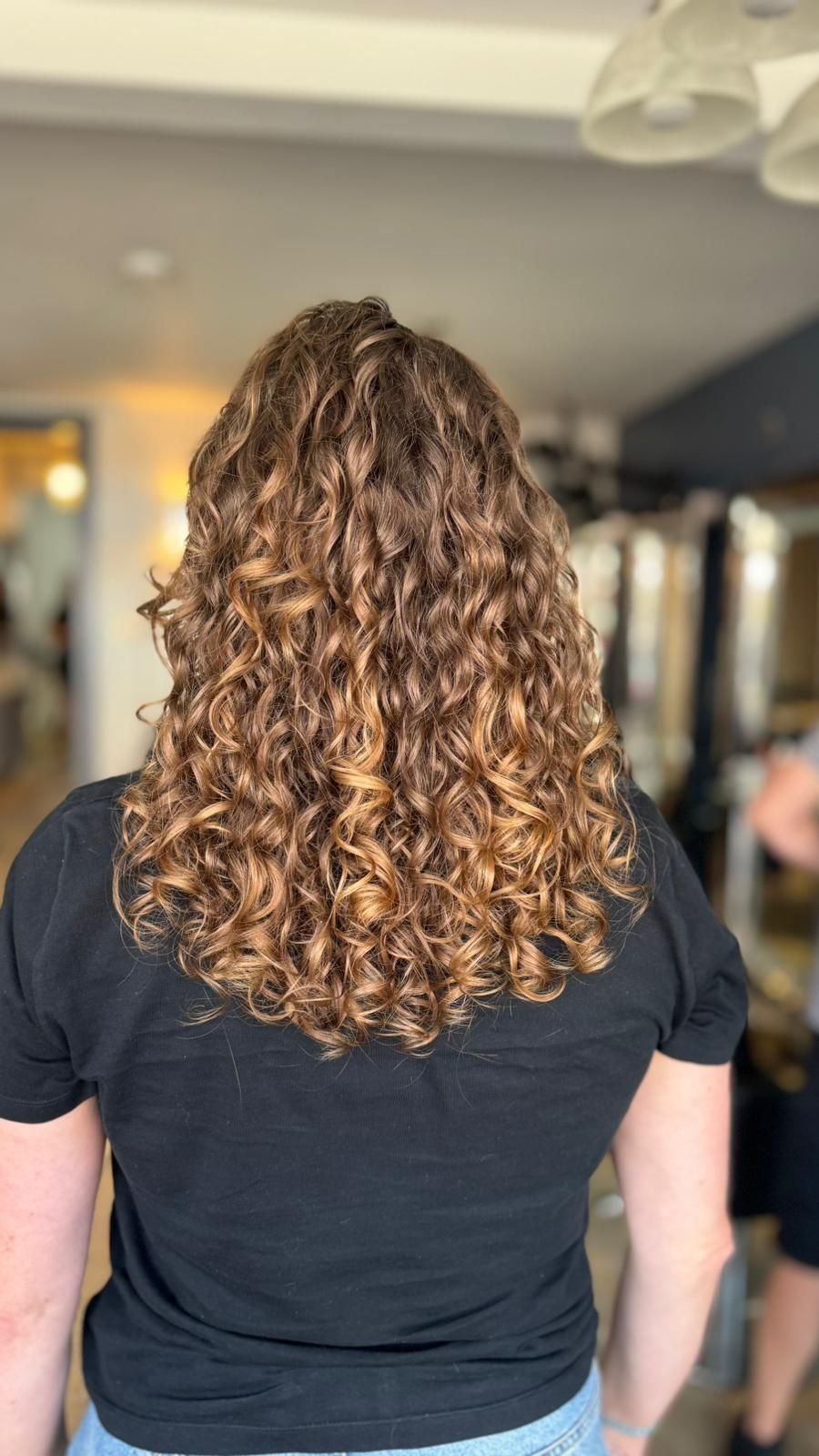 Back view of a person with curly brown hair wearing a black shirt in a salon interior