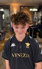 Person with curly hair in a black Venezia polo shirt, smiling in a barbershop chair