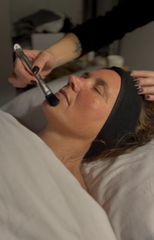 Facial treatment in progress as a brush is applied to a woman’s face while she lies on a bed