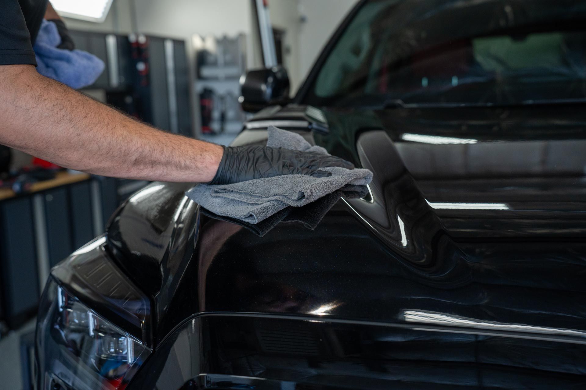 Person wearing gloves polishing a black car hood with a gray microfiber cloth in a garage.