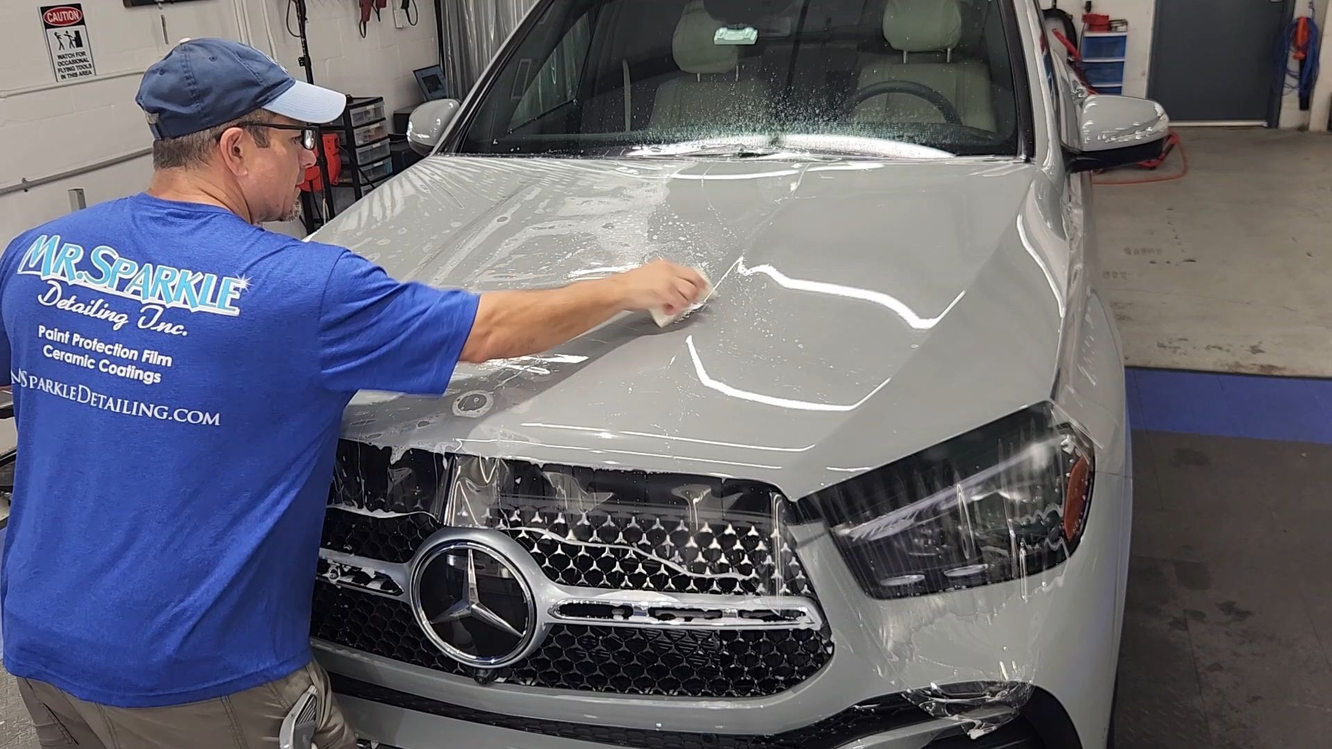 Man applying film to a white Mercedes car hood in a garage. He is wearing a blue shirt and hat.