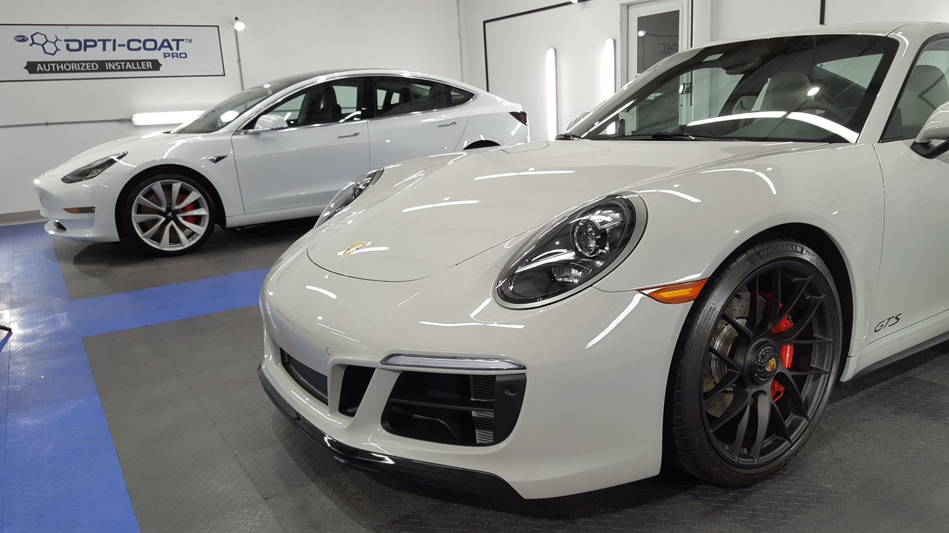 White Porsche 911 GTS and Tesla Model 3 inside a garage with bright lights, showcasing car detailing.