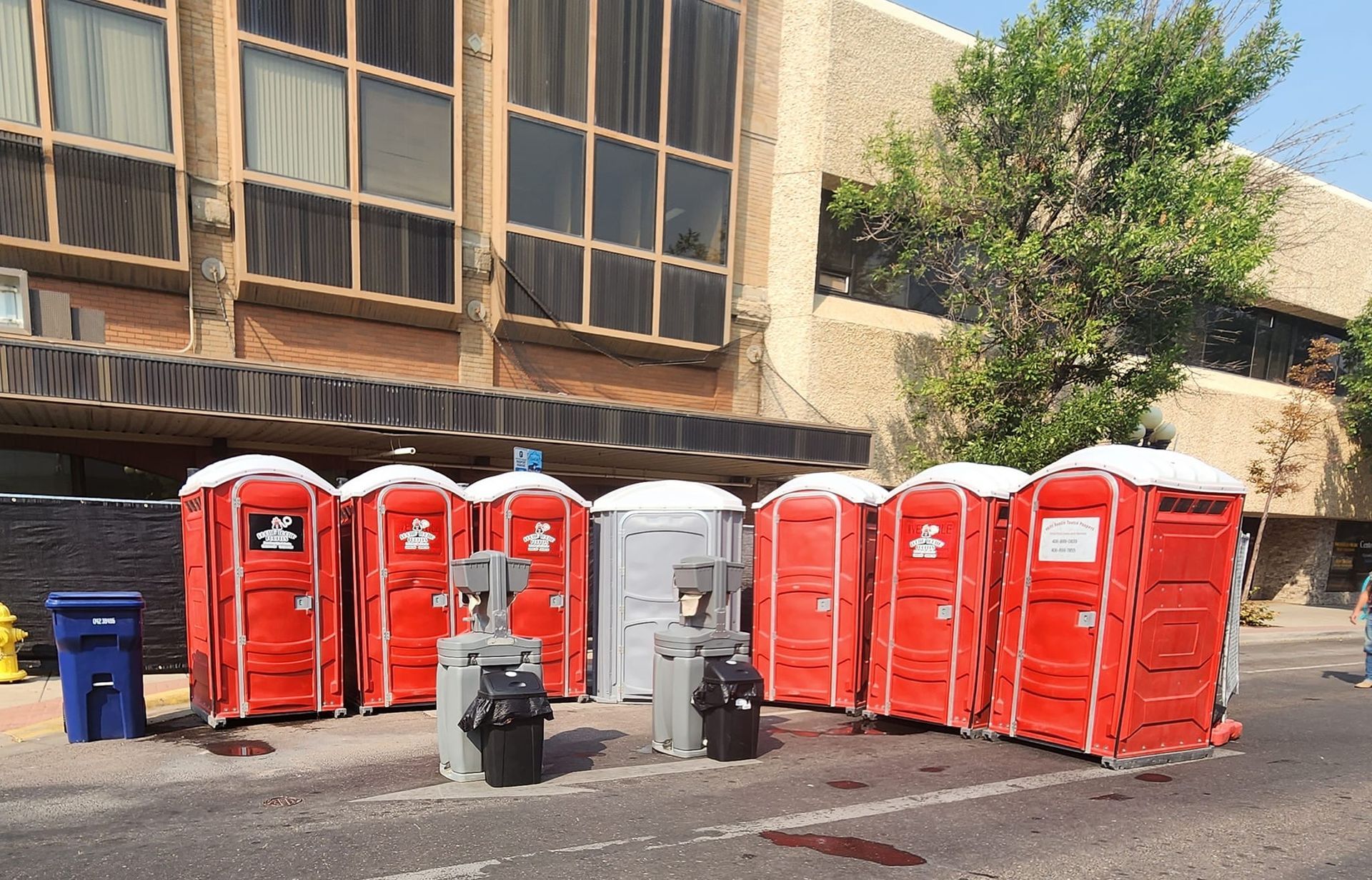 A row of red portable toilets and one gray unit placed on a street in front of a multi-story building.