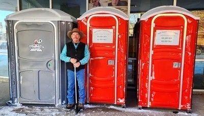 A person in a cowboy hat and vest stands holding a shovel between one grey and two red portable toilets.