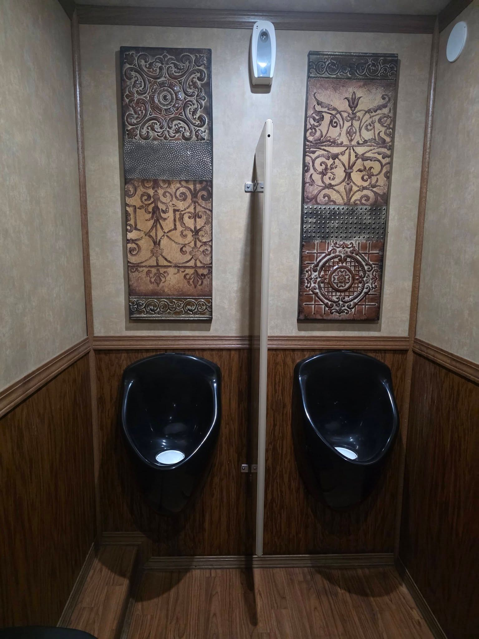 Two black wall-mounted urinals separated by a partition in a room with wood-paneled walls and decorative vertical art.
